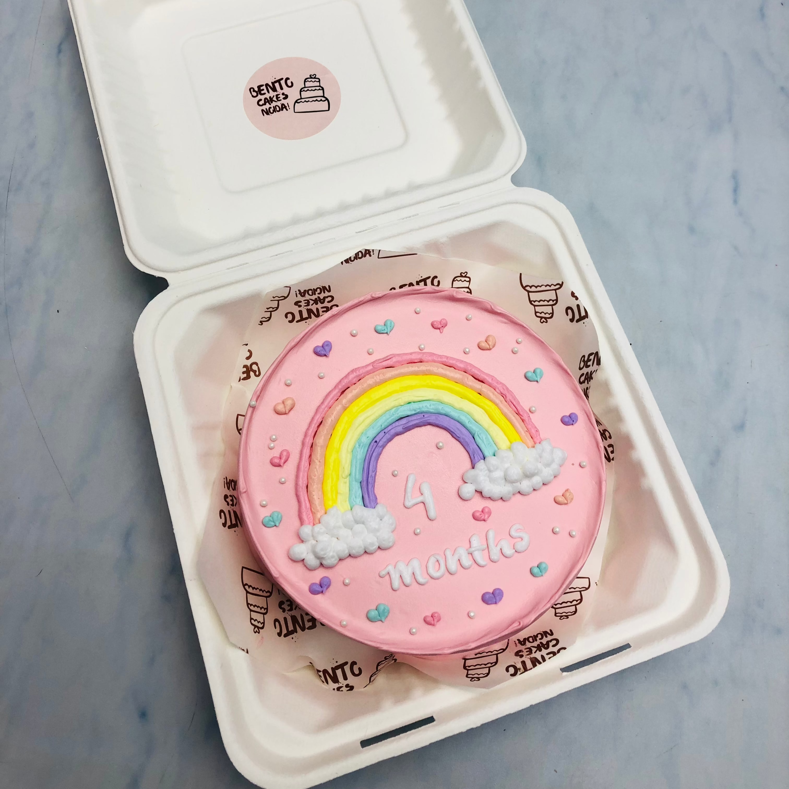 A light pink cake in a bento box designed with rainbow, clouds and prety colorful hearts all over the cake.