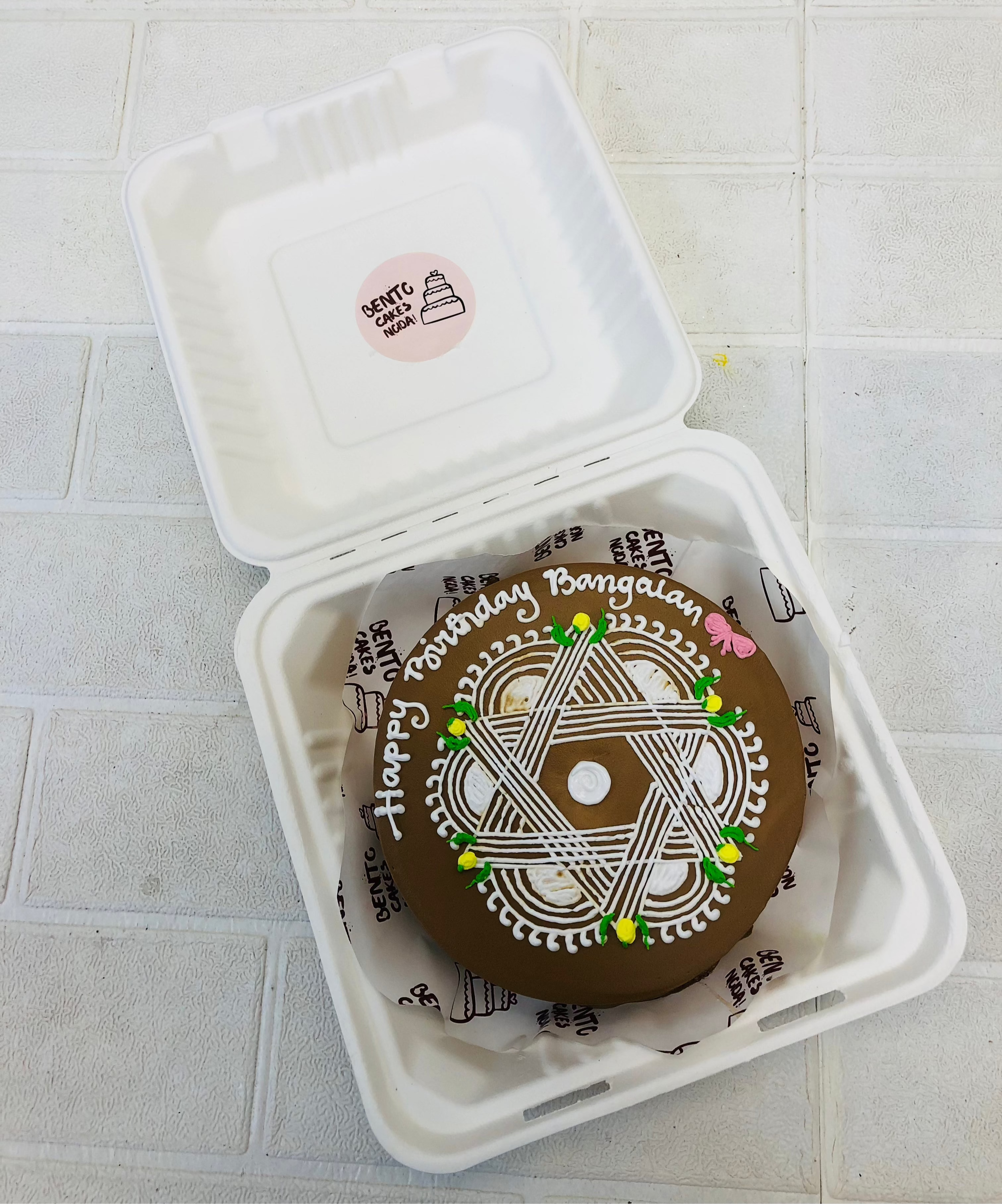 A beautifully decorated birthday cake inside a bento box. The cake features intricate white icing patterns with green and yellow accents, and the message "Happy Birthday Bangalan" written on top. A small pink butterfly adds a charming touch. The box is labeled "Crafty Cakes" with an illustration of a cake, enhancing its presentation.