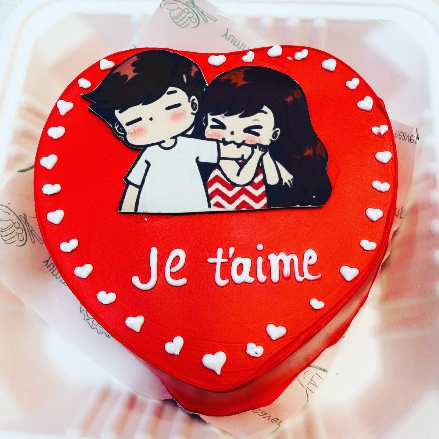 A red icing heart shape cake covered with white hearts on its surrounding and a biting couple sicker placed on the top of cake.