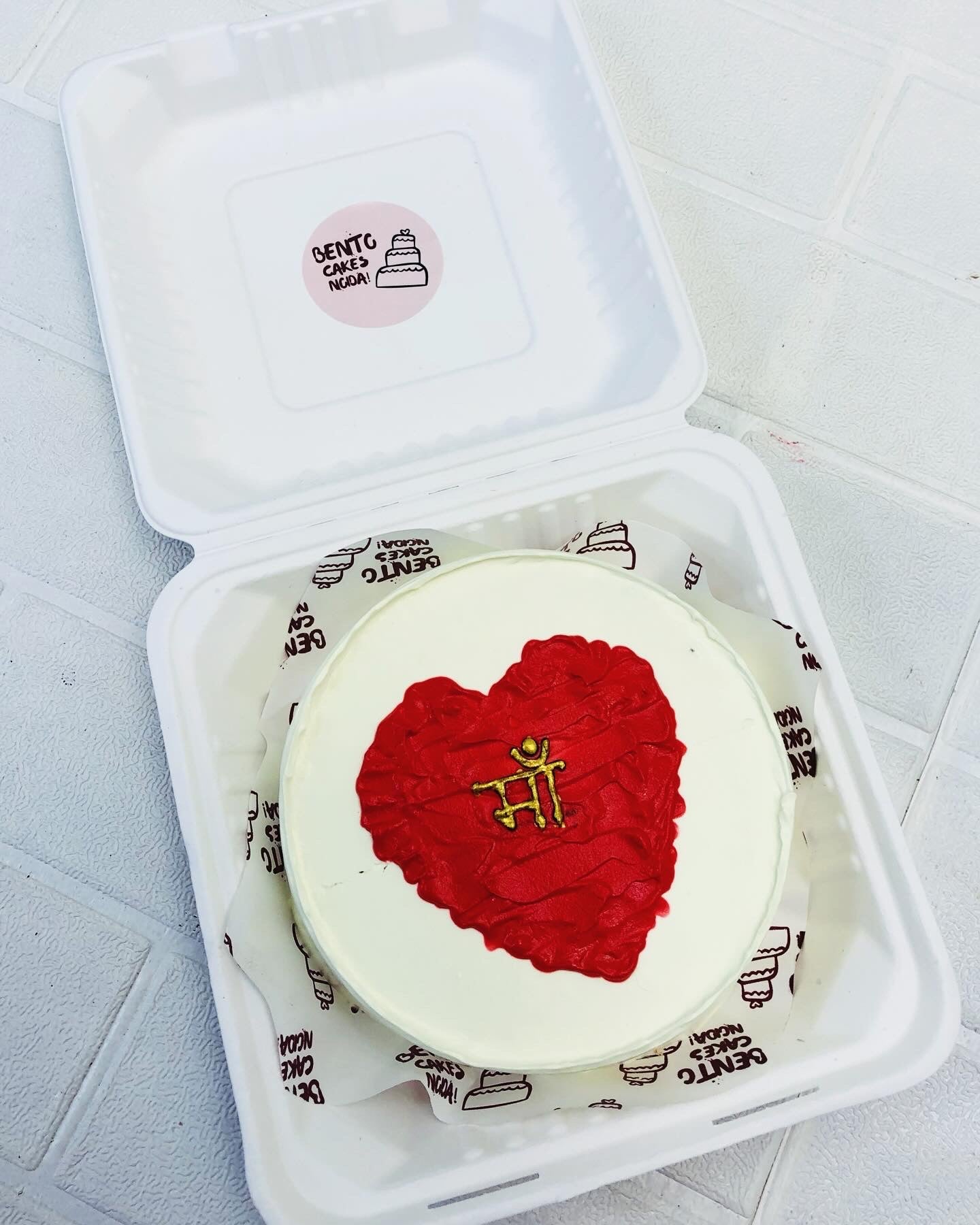 A white cake with dark red heart design in the center with a golden text off "Maa" on it.