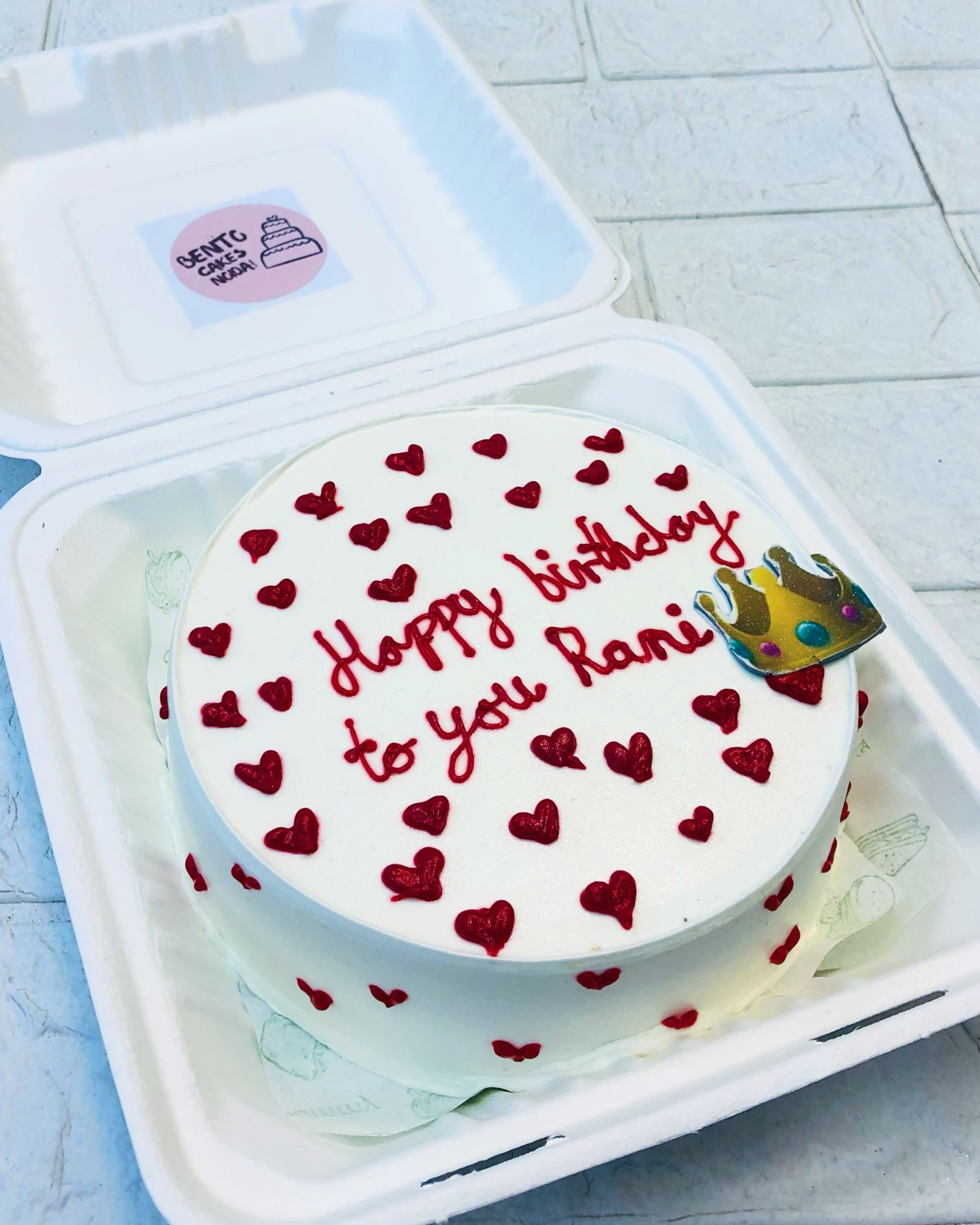 A white bento cake filled with creamy red hearts all ver the cake and a crown print on it with a text "Happy birthday to you Rani".