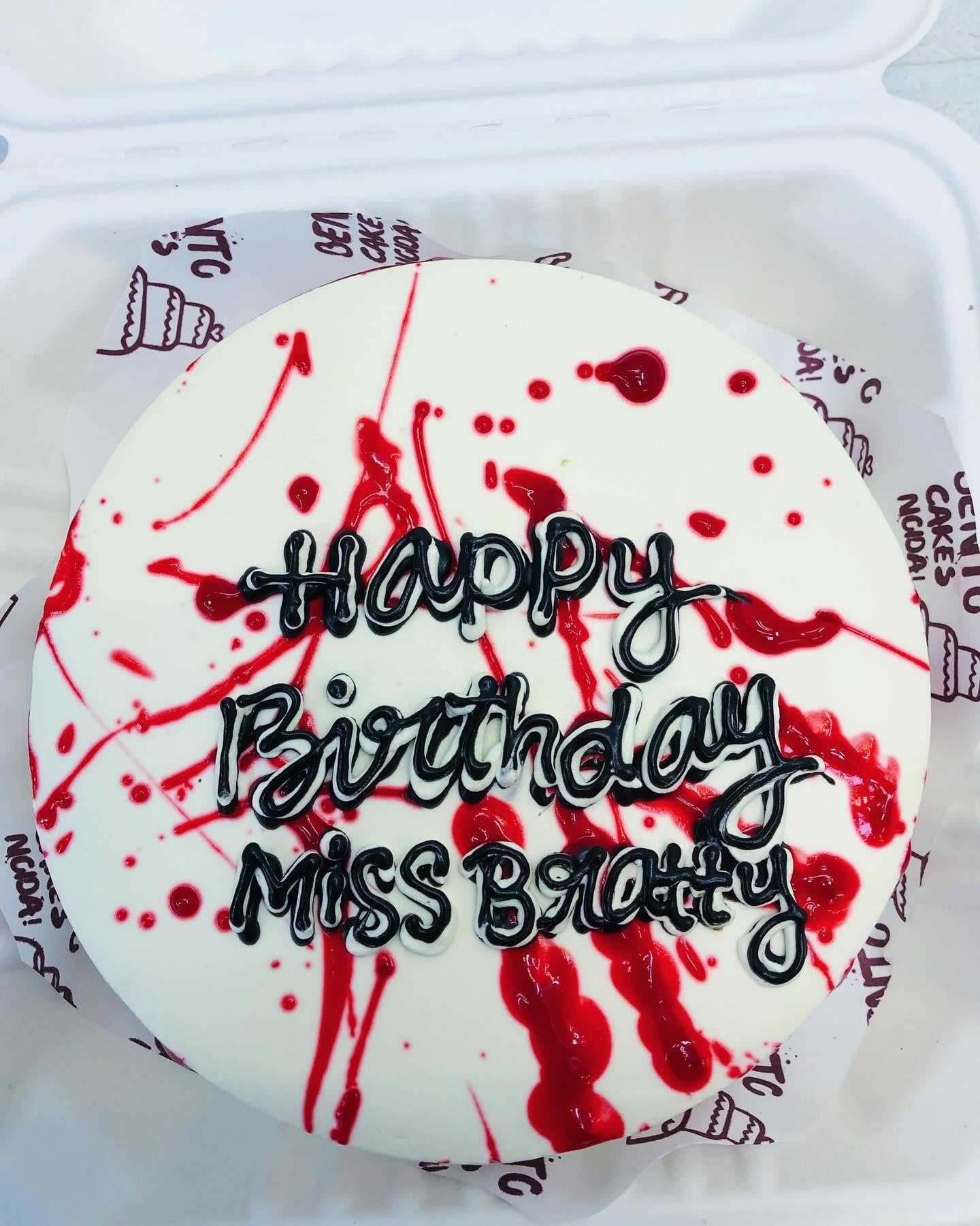 A uniquely designed birthday cake with white frosting, decorated with striking red splashes and black icing text that reads 'Happy Birthday Miss Bratty.'