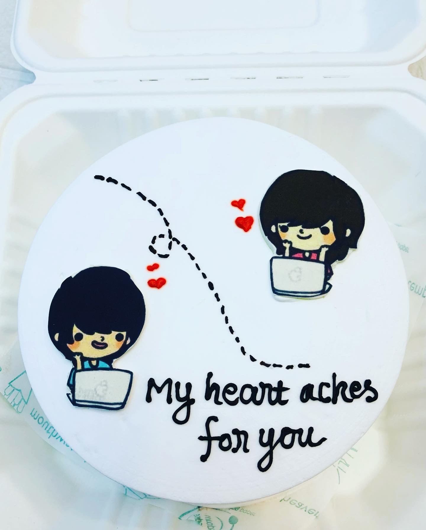 A round cake with a white frosting featuring two cartoon characters using laptops, connected by a dotted line with red hearts, and the text 'My heart aches for you'.