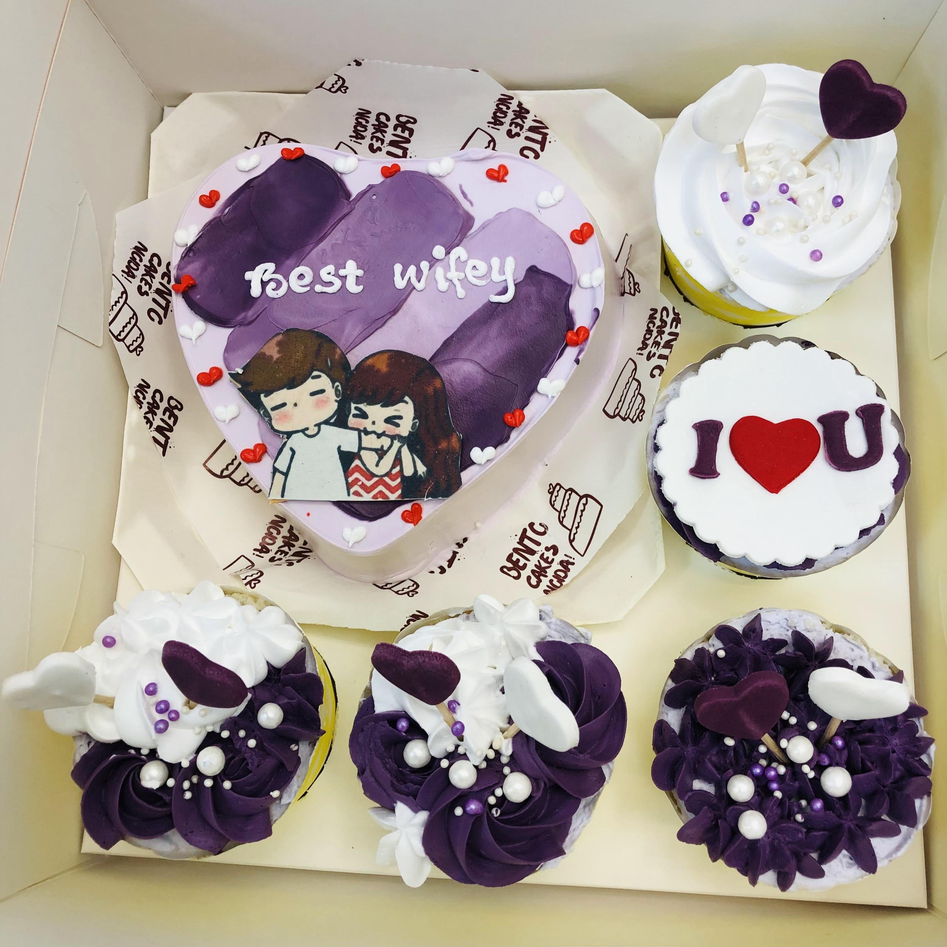 A heart-shaped cake with the message "Best Wifey" and an illustration of a couple, decorated with small red and white heart-shaped toppings. Surrounding the cake are four cupcakes with various designs, including one with "I ♥ U" and others decorated with purple and white frosting, heart toppers, and edible pearls, all presented in a white bento box labeled "Bento Cakes Korea."