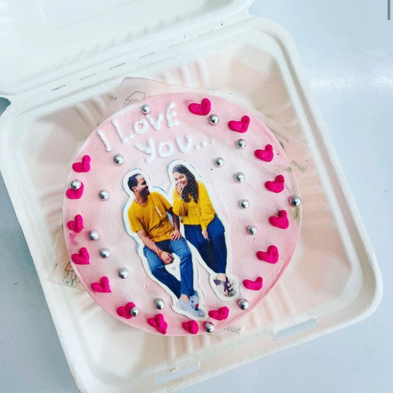 A round cake with pink frosting, decorated with a photo of a couple wearing yellow tops and blue jeans. The cake is adorned with pink heart-shaped decorations and silver edible balls. The text "I LOVE YOU..." is written on the top of the cake in white icing.