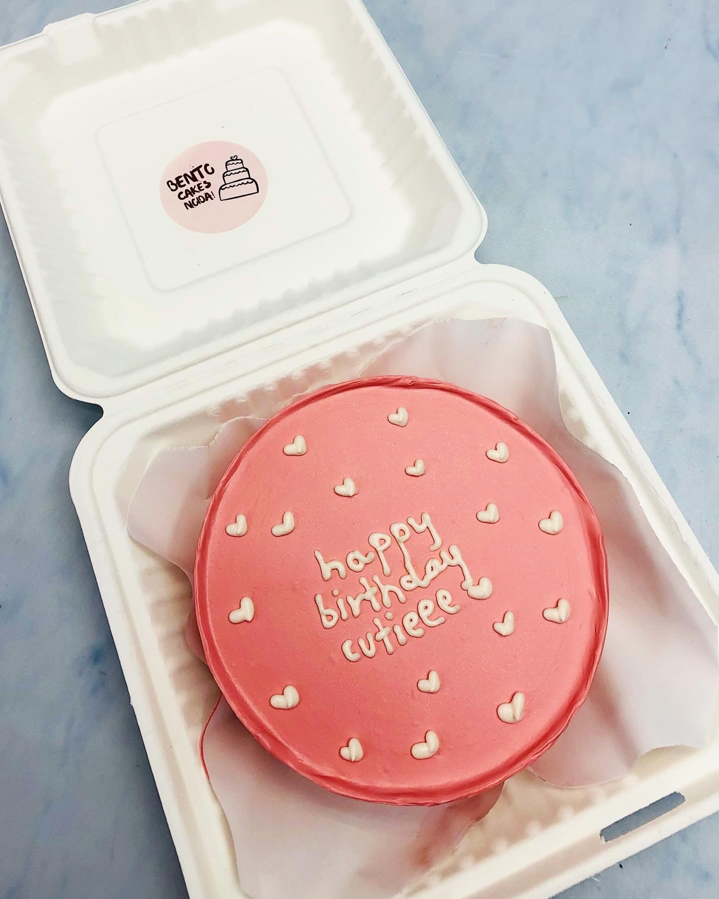 A rose gold bento cake with white hearts design and a text written with white icing.