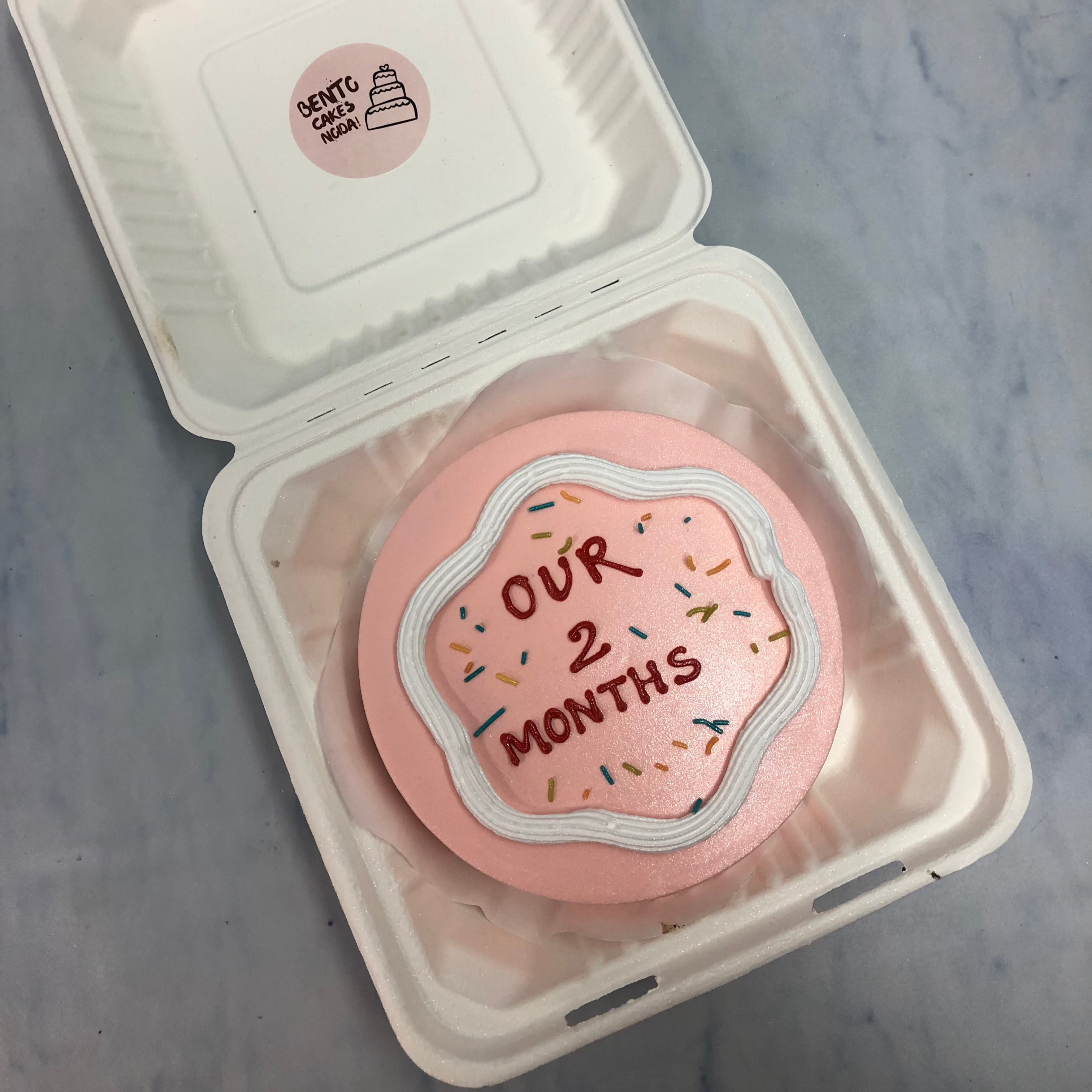 A rose gold bento cake with white creamy design and loving text on it.
