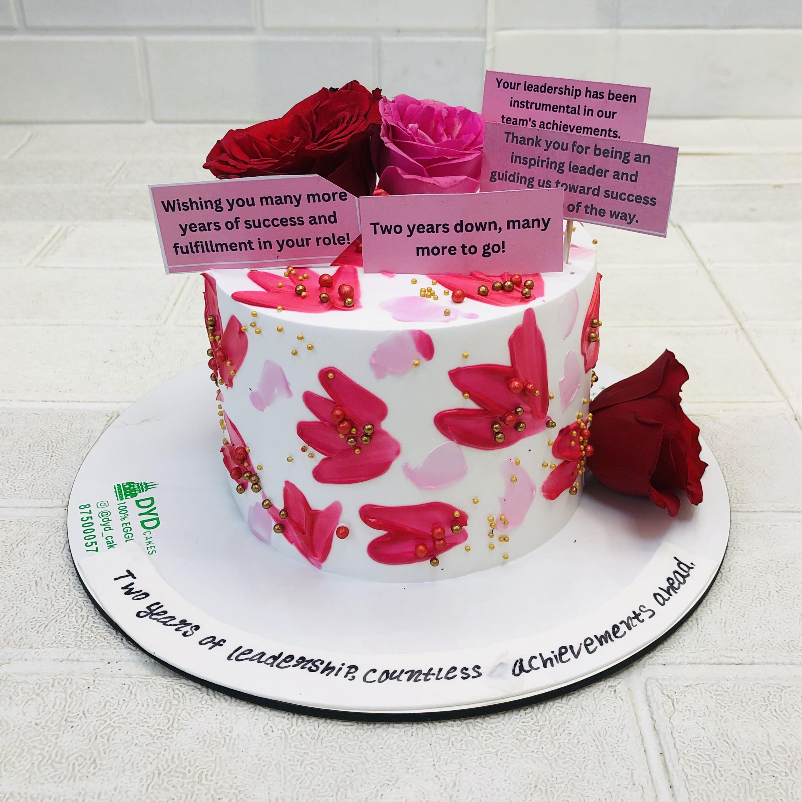 A pretty white pink cake decorated with red and pink roses on its top and bottom with some message stickers and surrounded by flowers patterns on it.