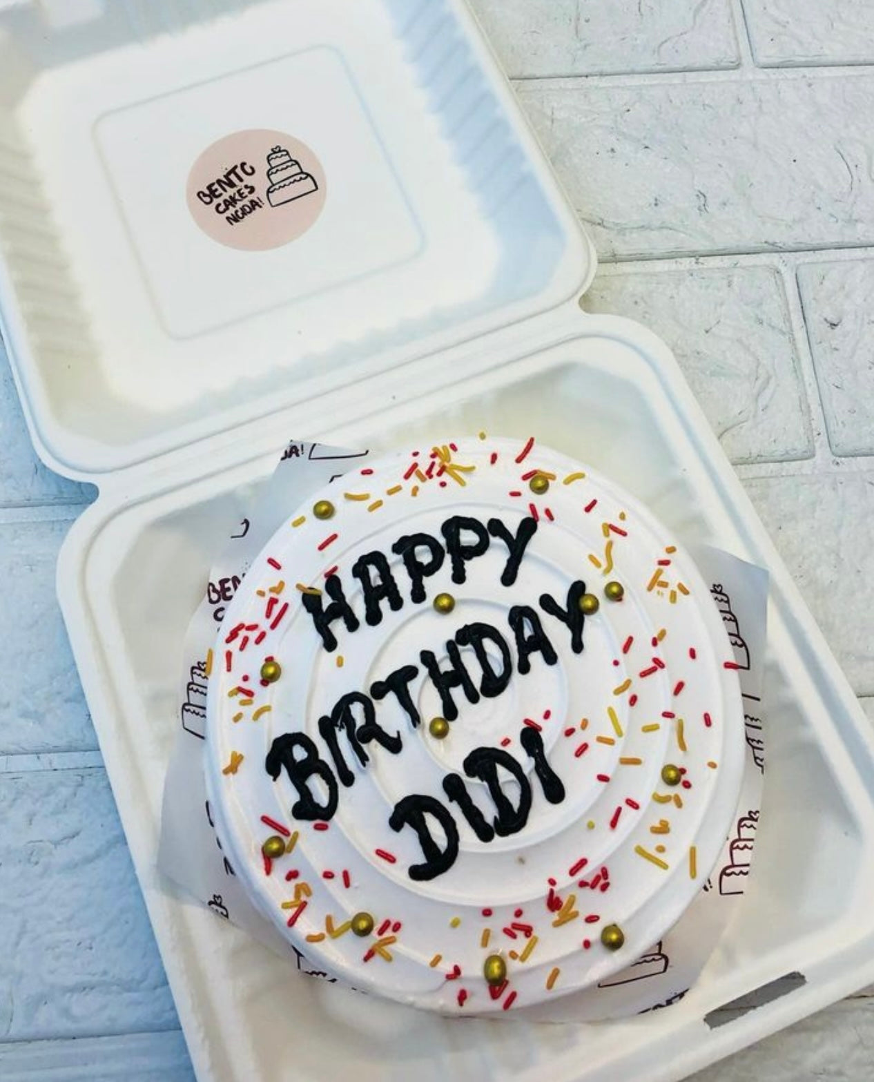 A neatly presented white frosted bento cake with the message "HAPPY BIRTHDAY DIDI" in black icing, adorned with red and yellow sprinkles and golden beads, placed inside a bento box.