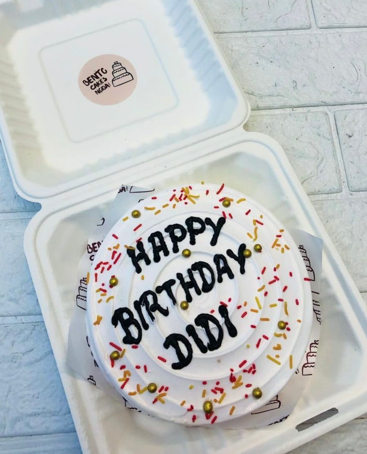 A neatly presented white frosted bento cake with the message "HAPPY BIRTHDAY DIDI" in black icing, adorned with red and yellow sprinkles and golden beads, placed inside a bento box.