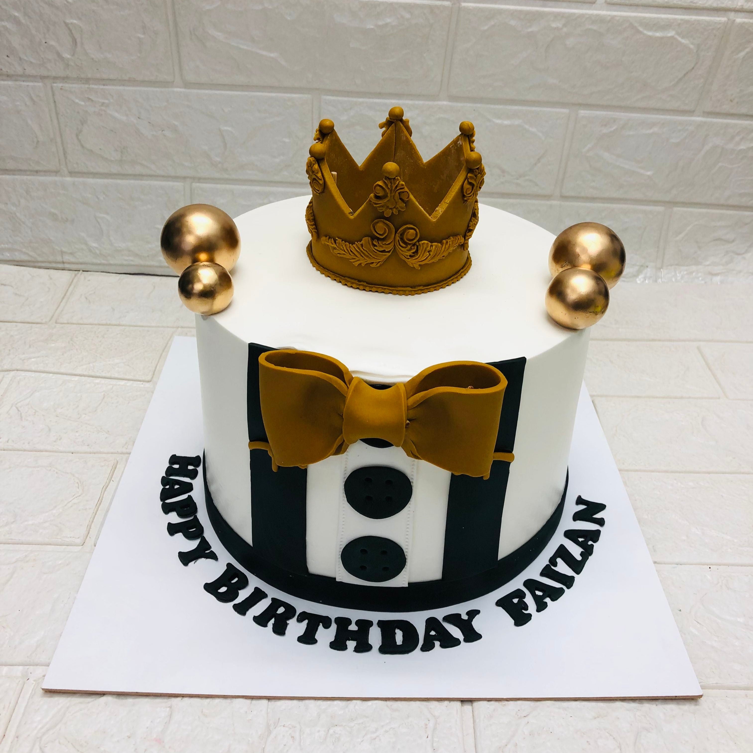 A white birthday cake adorned with a golden crown, golden bow tie, and black buttons, topped with two golden spheres. The cake board reads "HAPPY BIRTHDAY FAIZAN" in black letters.