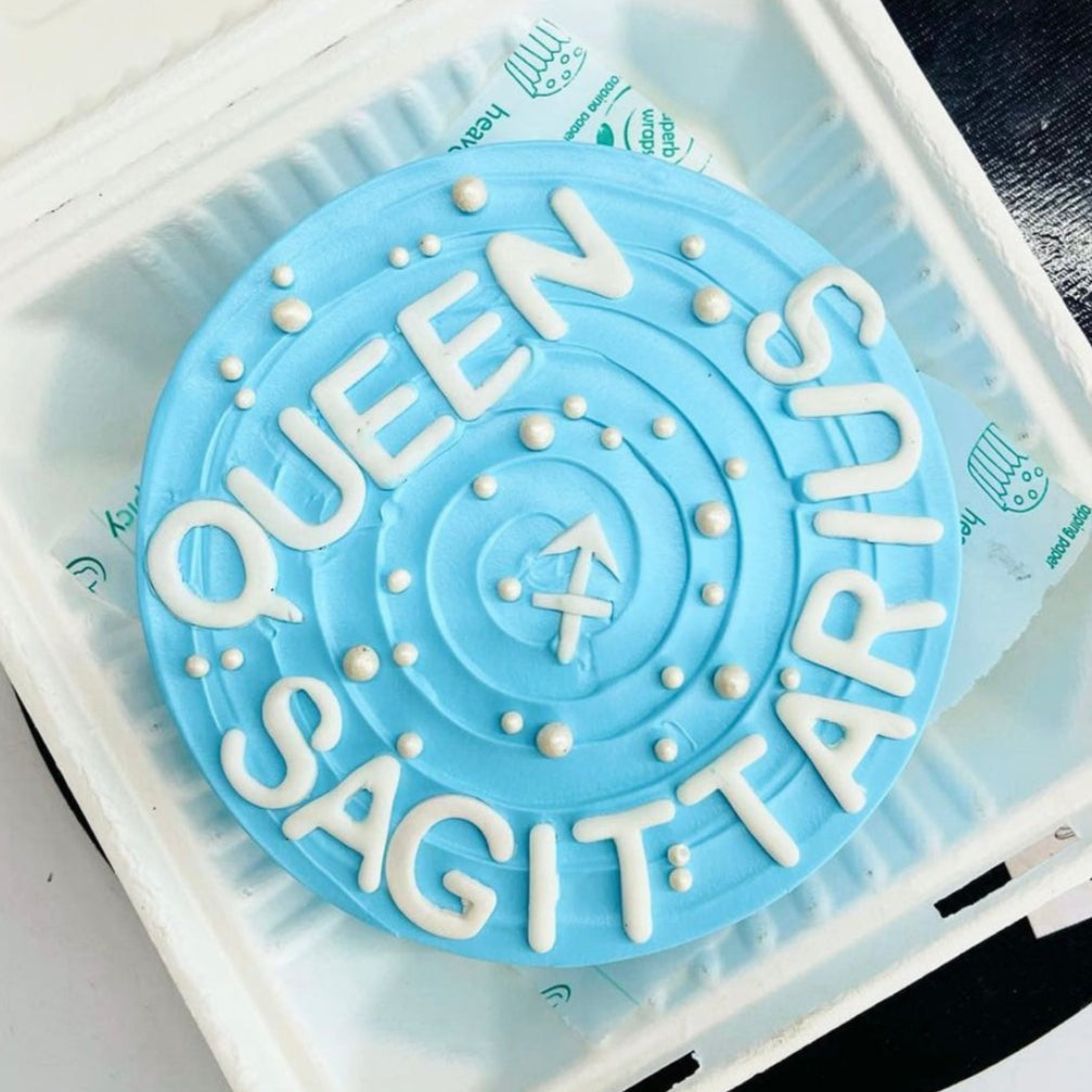 A beautifully decorated bento cake featuring the phrase "Queen Sagittarius" on its creamy top.