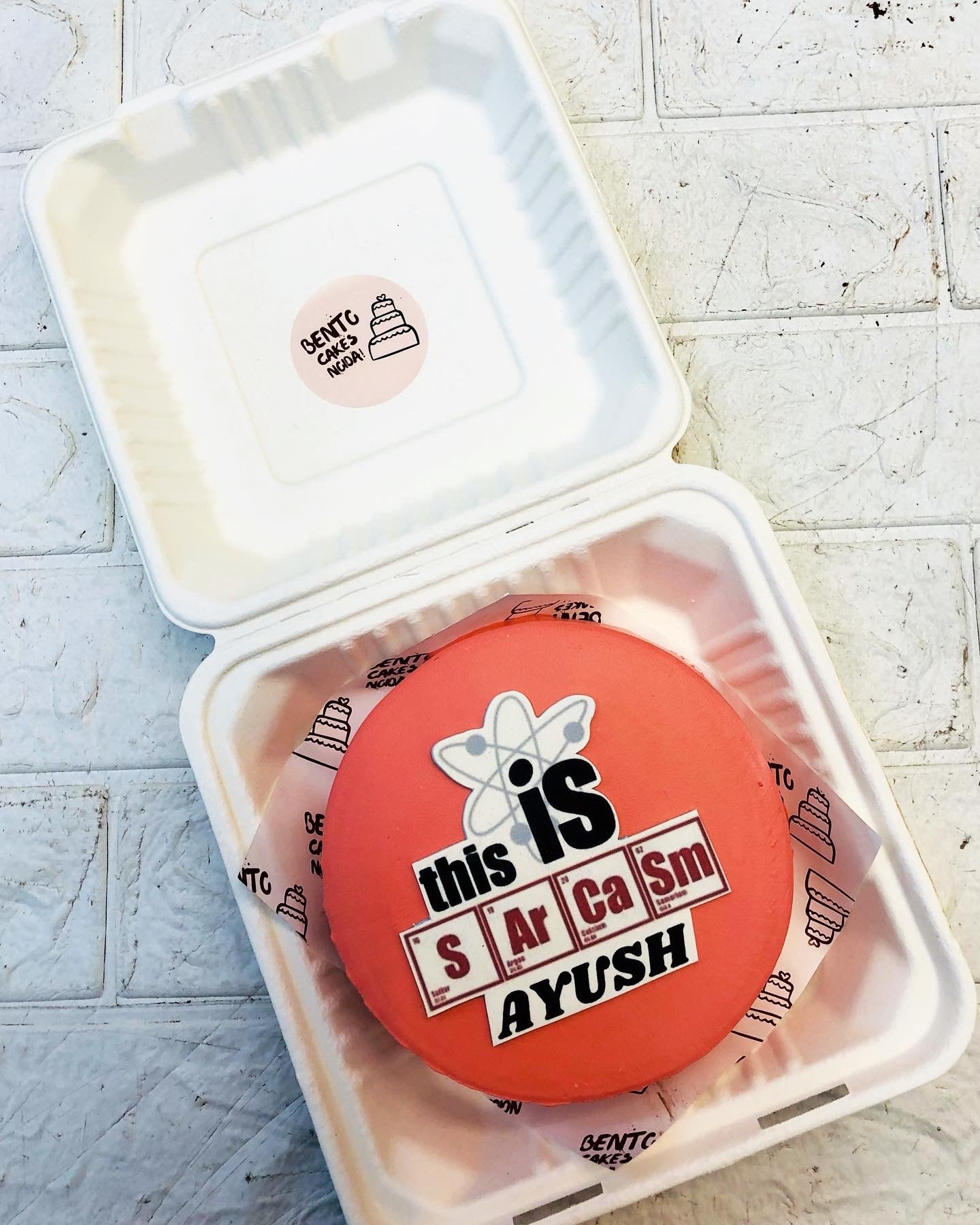 A round, red bento cake placed inside a white container, featuring a humorous printed message that reads, "this is sarcasm AYUSH" with a stylized atom symbol.