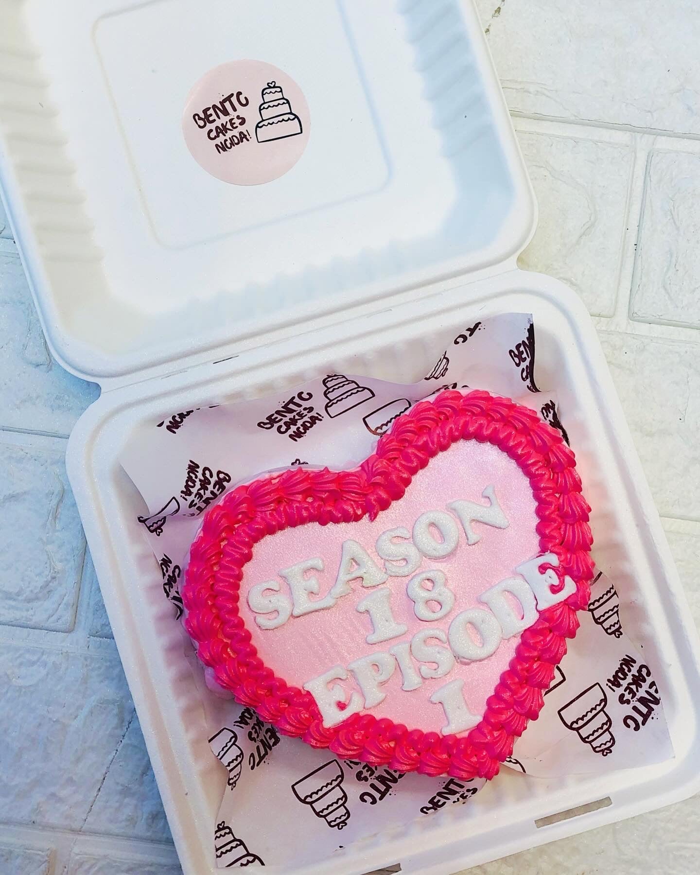 A light pink heart shape cake with dark pink design on it's boundary and a text with white fondant letters written on it.
