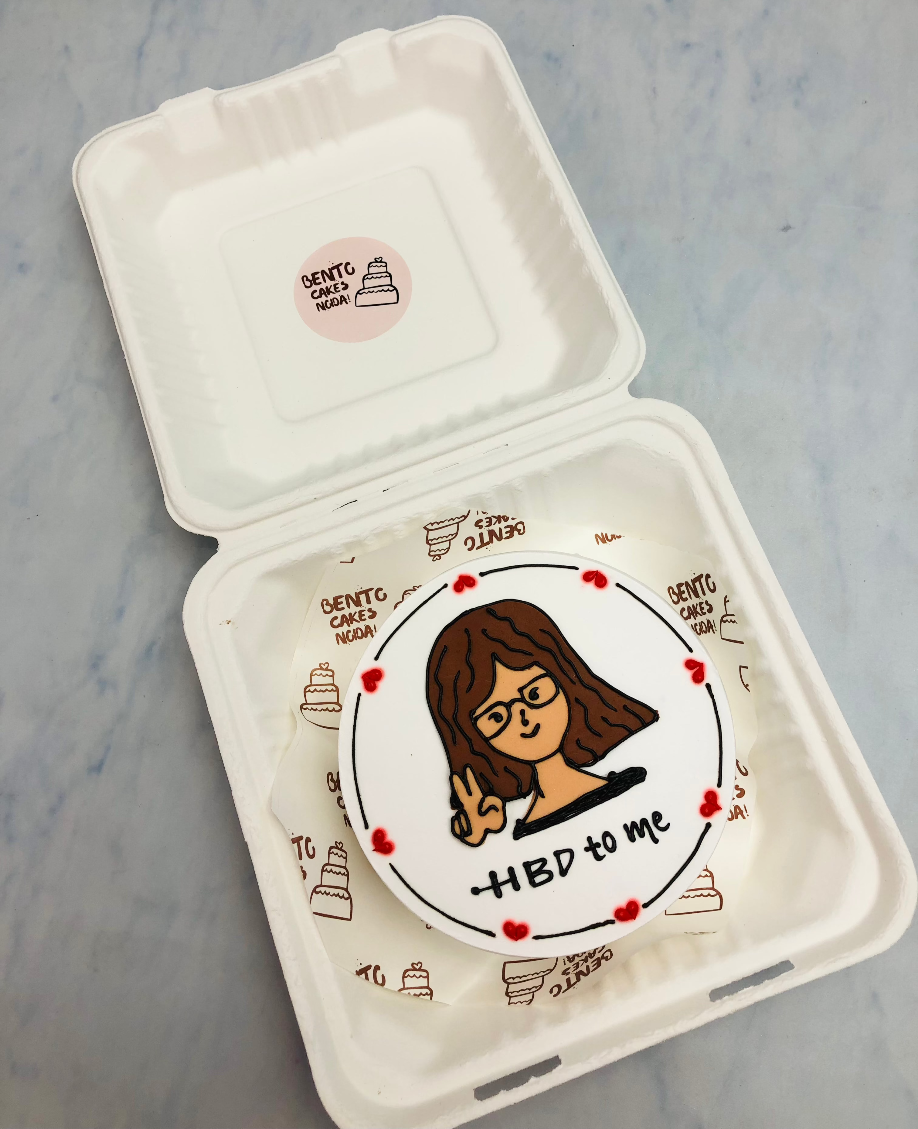 A compact, round bento cake featuring a hand-drawn cartoon portrait of a person wearing glasses and making a peace sign. The cake is decorated with red dots along the edge and has "HBD to me" written on it.