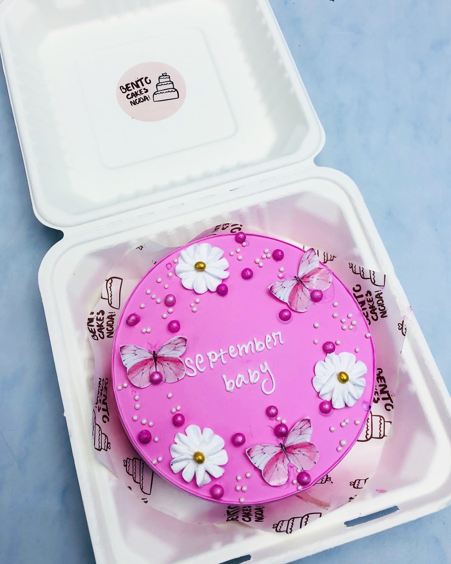 A pink birthday cake with the text 'September baby' written on it, decorated with white flowers, pink butterflies, and small pearls. 