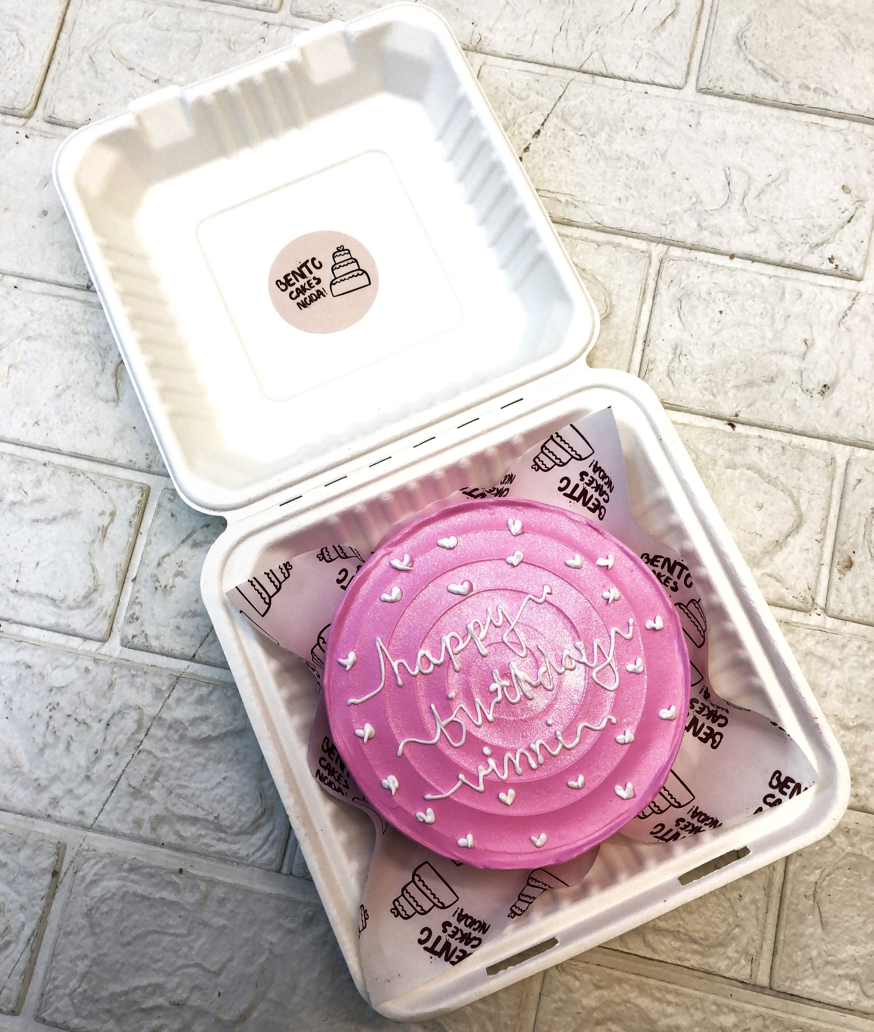 A shiny pink bento cake decorated with white hearts design and and a text "happy birthday vinni" written with white icing on it.