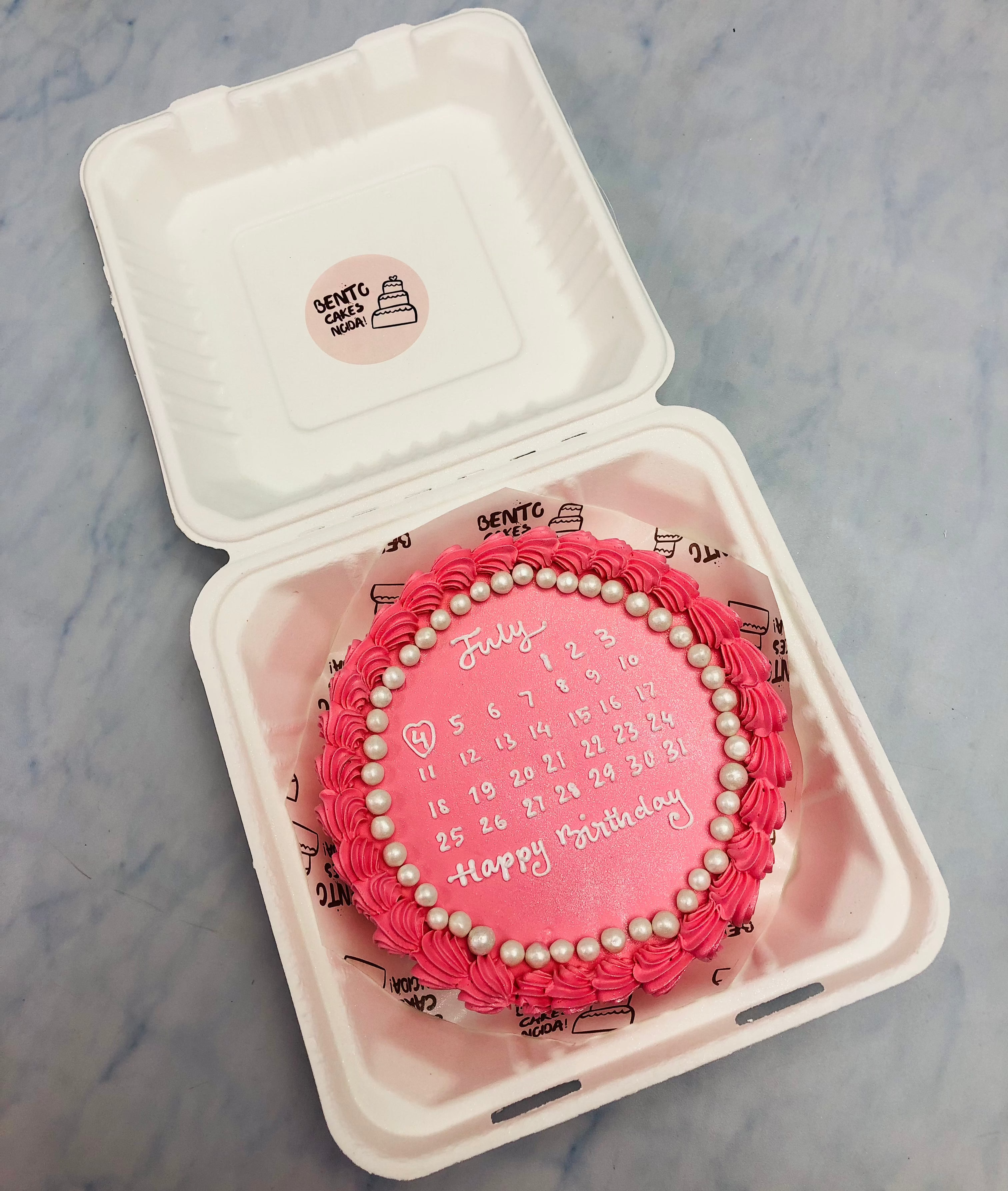 A shiny pink bento cake having white calendar design and decorated with white sprinkles.