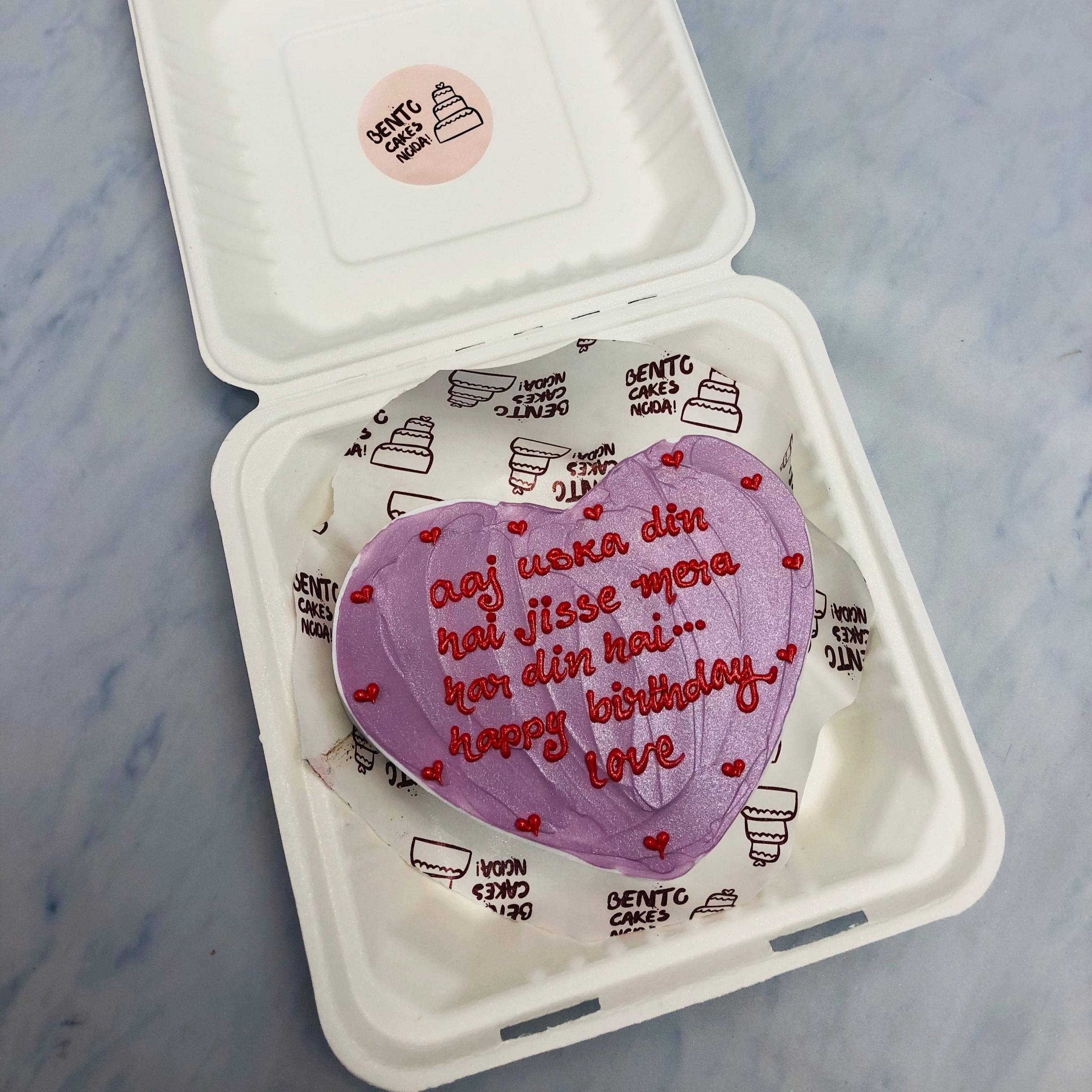 A white heart shape cake with shiney purple icing on its top and a loving text writtren on it with red icing.