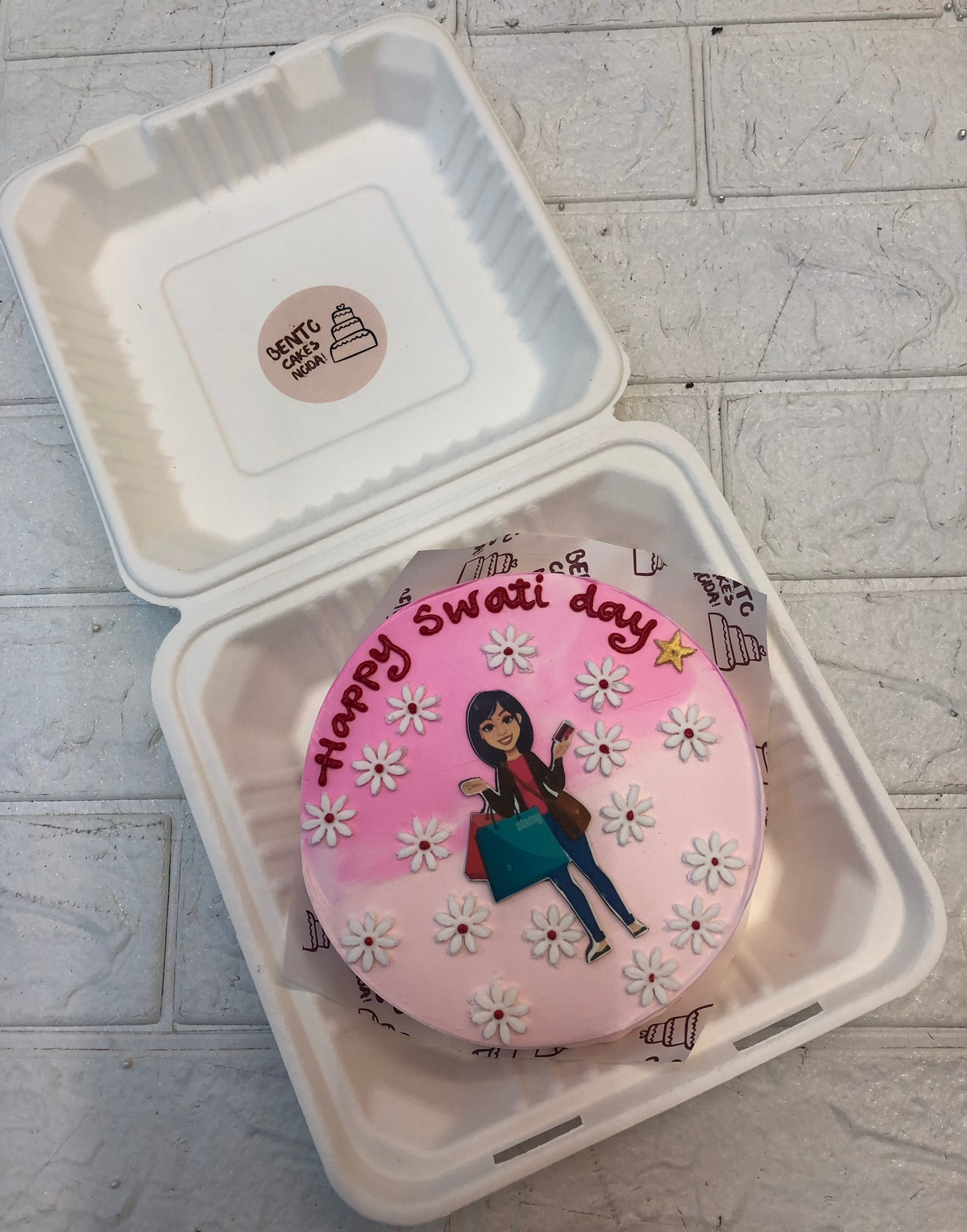 A round pink bento cake with white floral decorations and a hand-drawn figure holding a bag and a phone, placed inside a white bento box. The cake features 'Happy Swati day' written in red icing.
