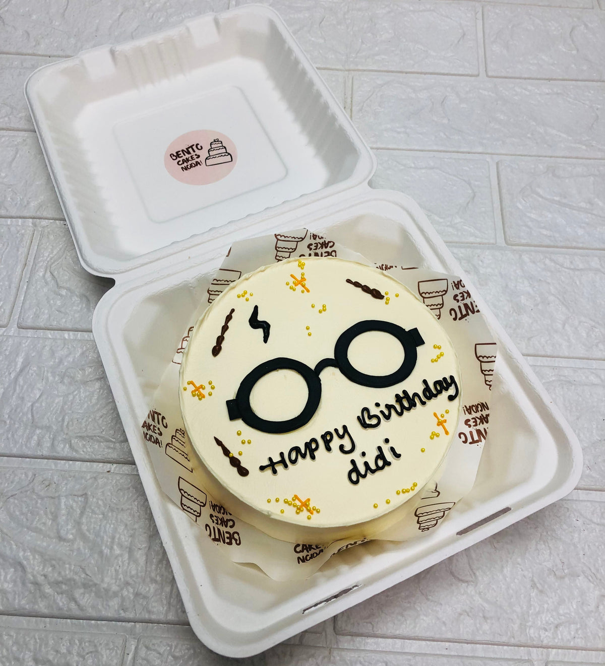 An offwhite bento cake having a goggle design and decorated with golden sprinkles.