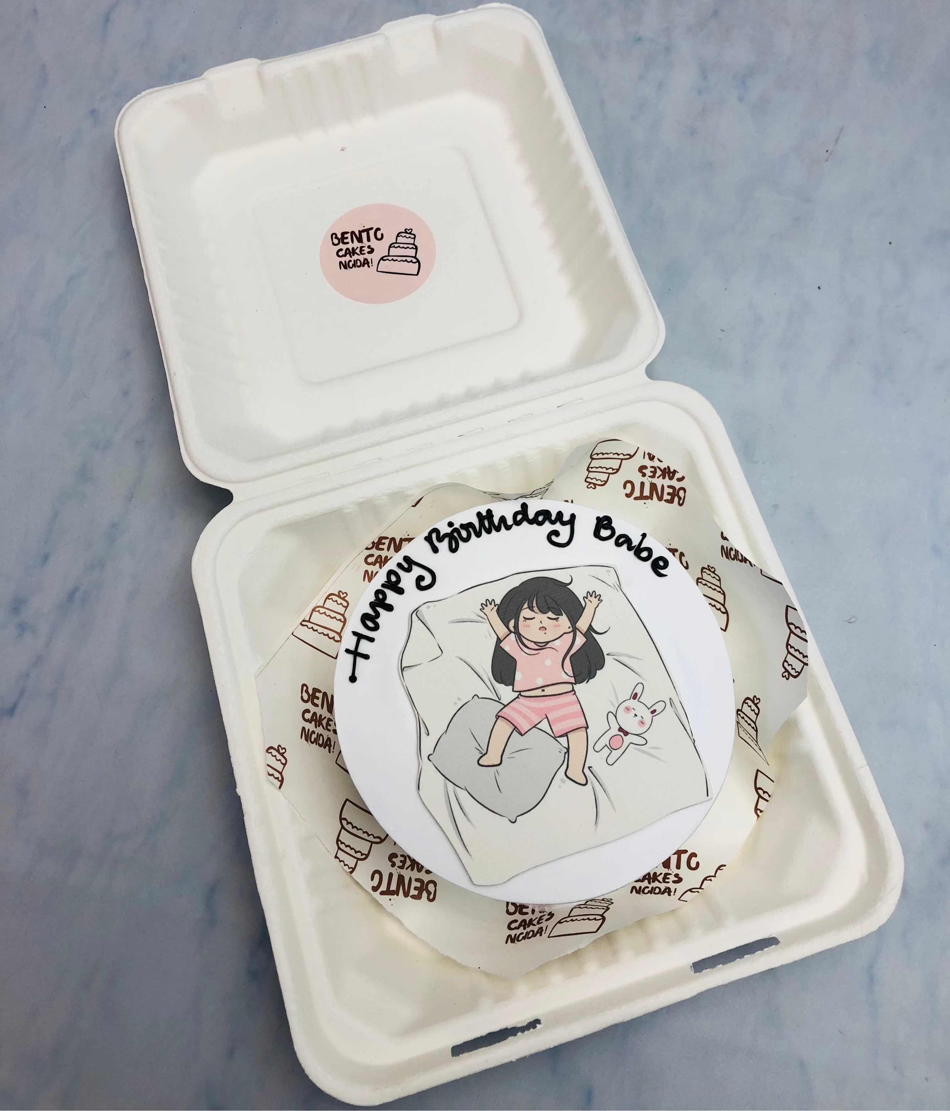The cake features a custom illustration of a girl in pink pajamas lying on a bed with a white bunny. The top of the cake has the text "Happy birthday Babe" written in black icing.