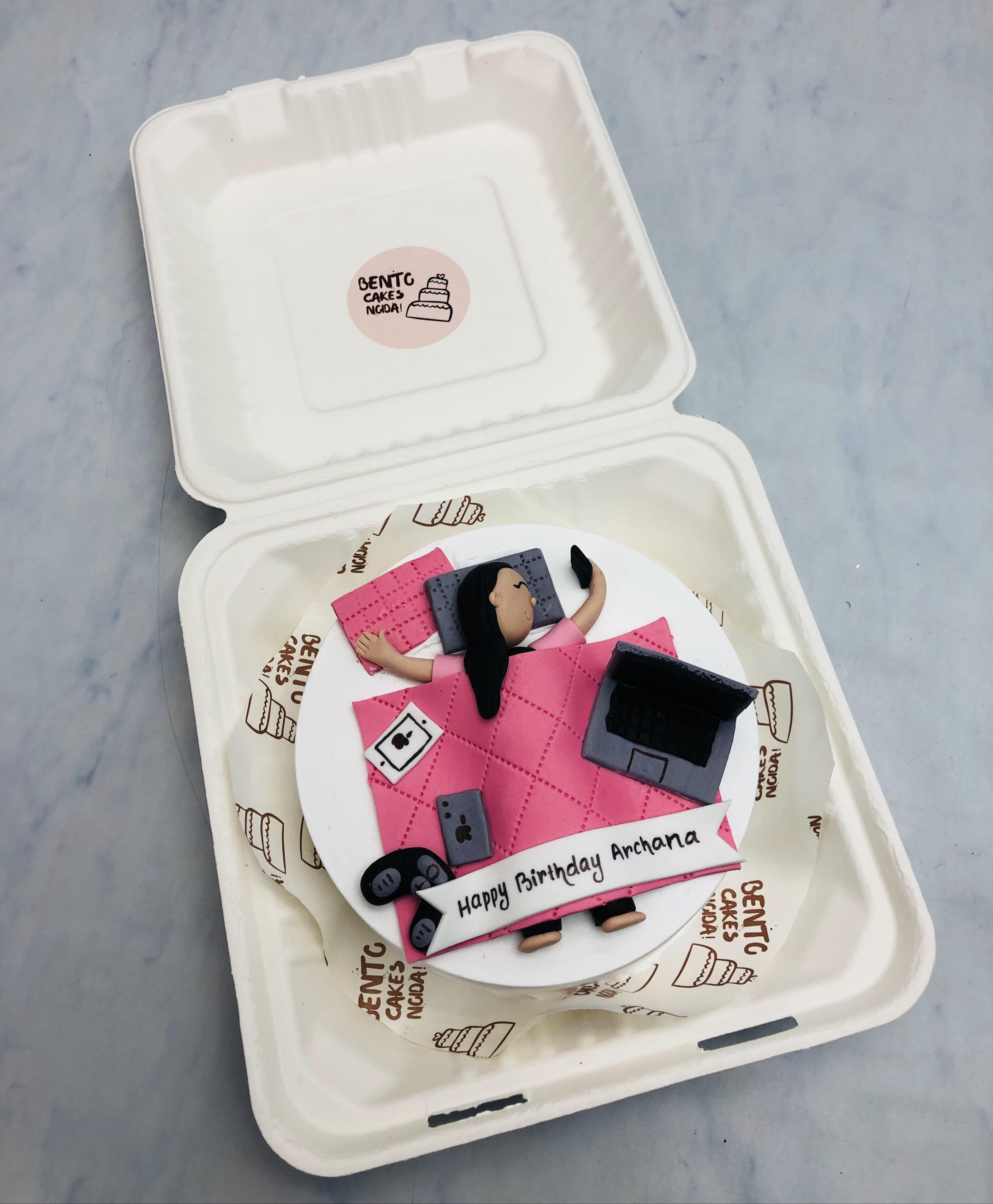 A personalized birthday cake inside a white takeout box, decorated with a fondant figure lying on a pink quilted blanket, surrounded by a laptop, smartphone, and headphones. The cake features the message "Happy Birthday Archana".