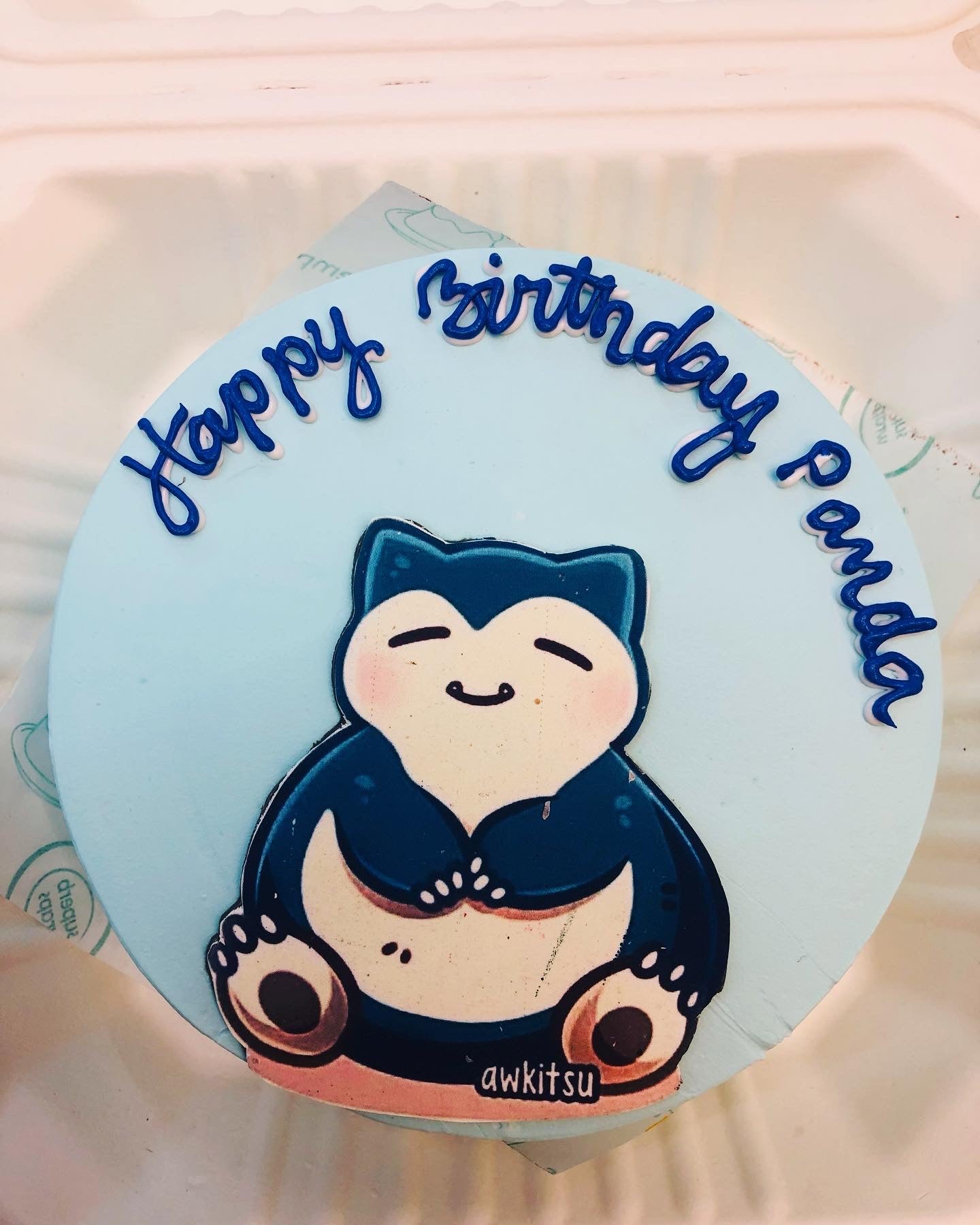 A round birthday cake with light blue frosting featuring a cartoon character, Snorlax, from Pokémon. The cake has the text "Happy Birthday Paula" written in blue icing around the top edge.