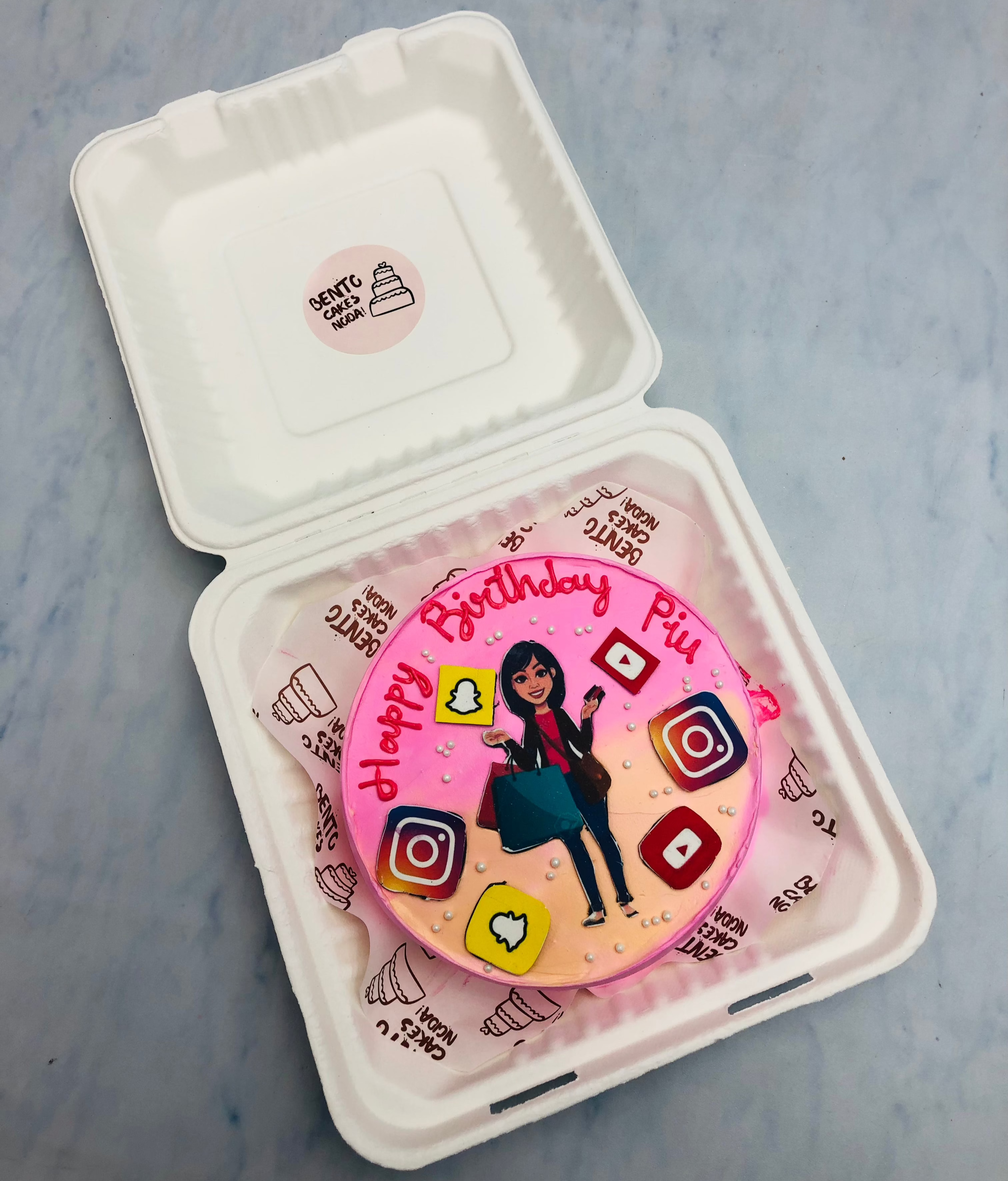 A uniquely designed birthday Bento cake with a pink and purple gradient background, decorated with social media icons (Instagram, Snapchat, YouTube) and a businesswoman illustration, placed inside a bento box.