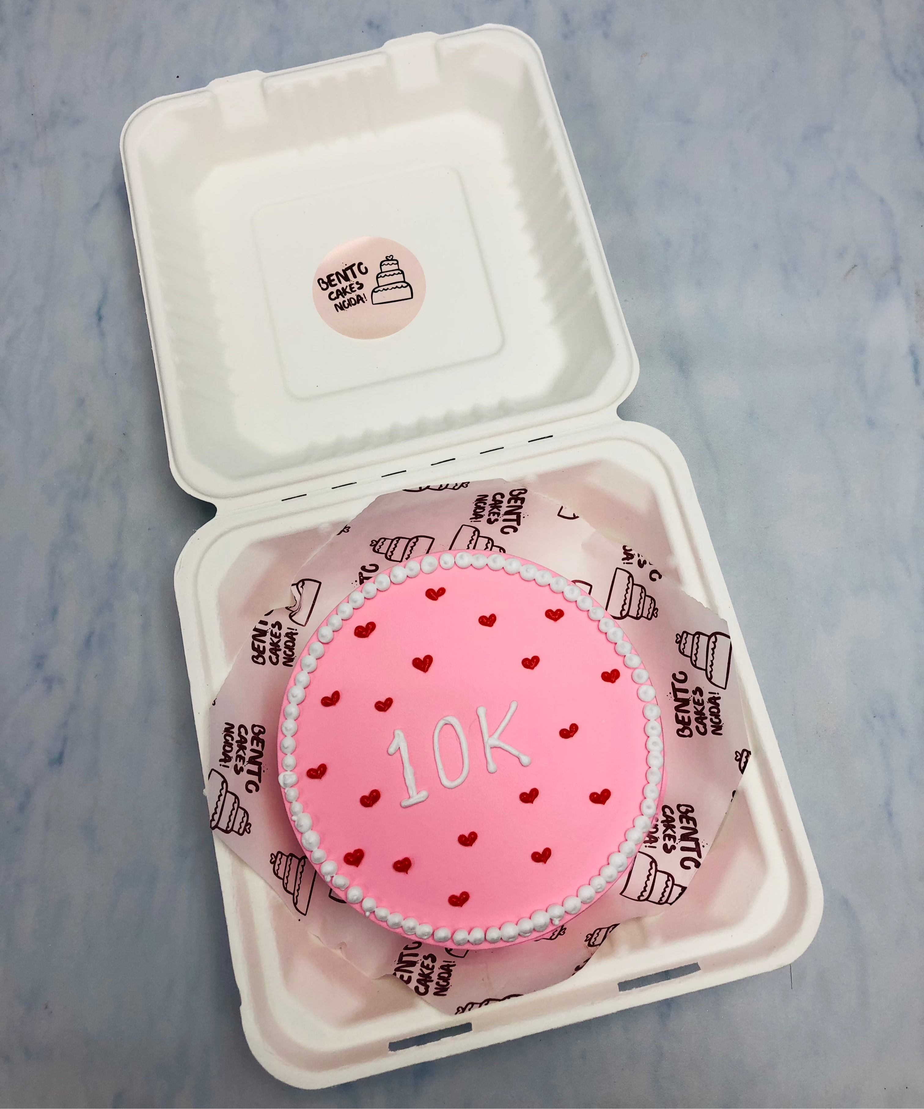 A light pink bento cake is decorated with little red hearts and a 10K text written with white icing.