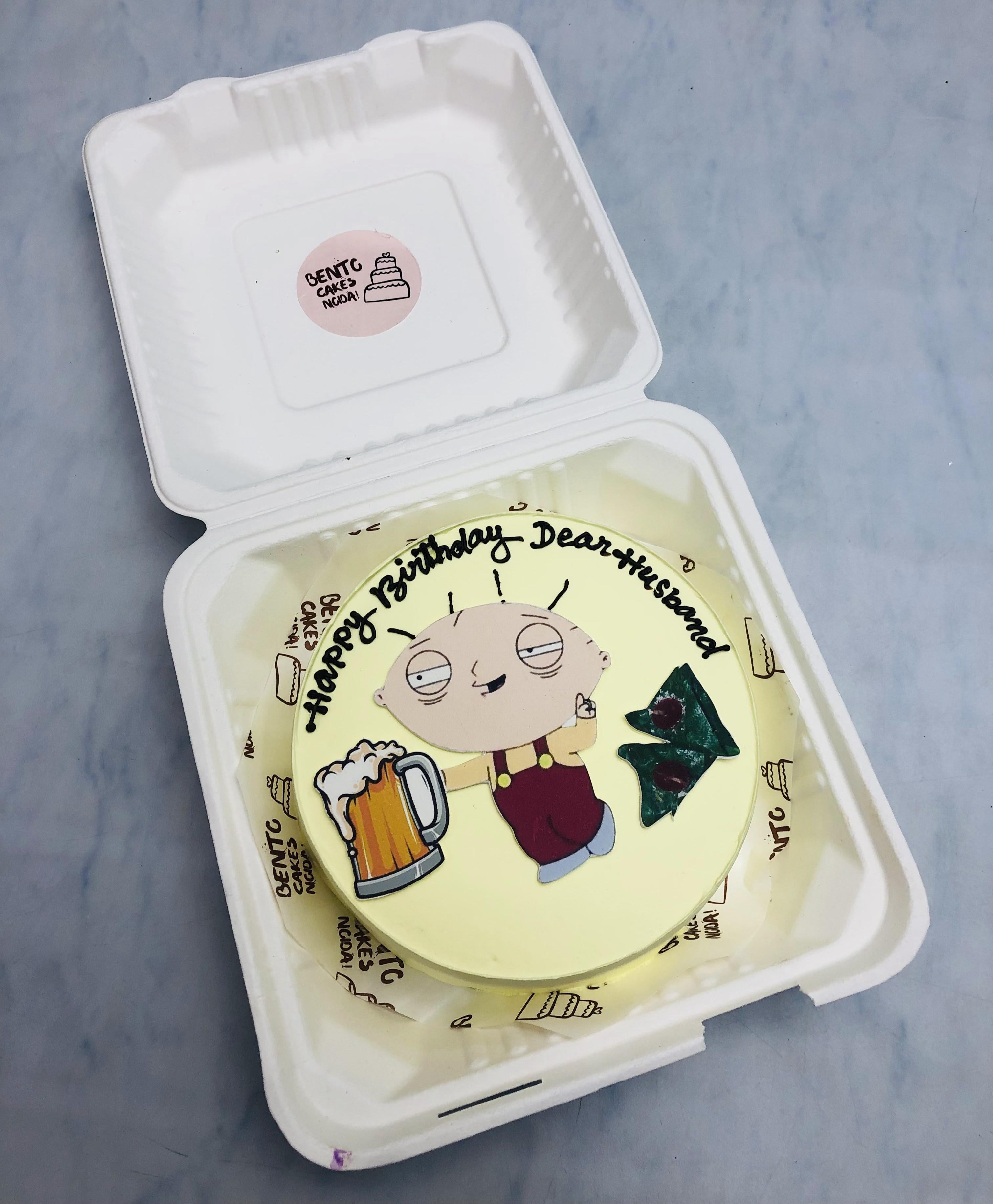 A birthday cake featuring Stewie Griffin from Family Guy, holding a beer mug, with the text "Happy Birthday Dear Husband" written on it. The cake is placed inside a white bento box.