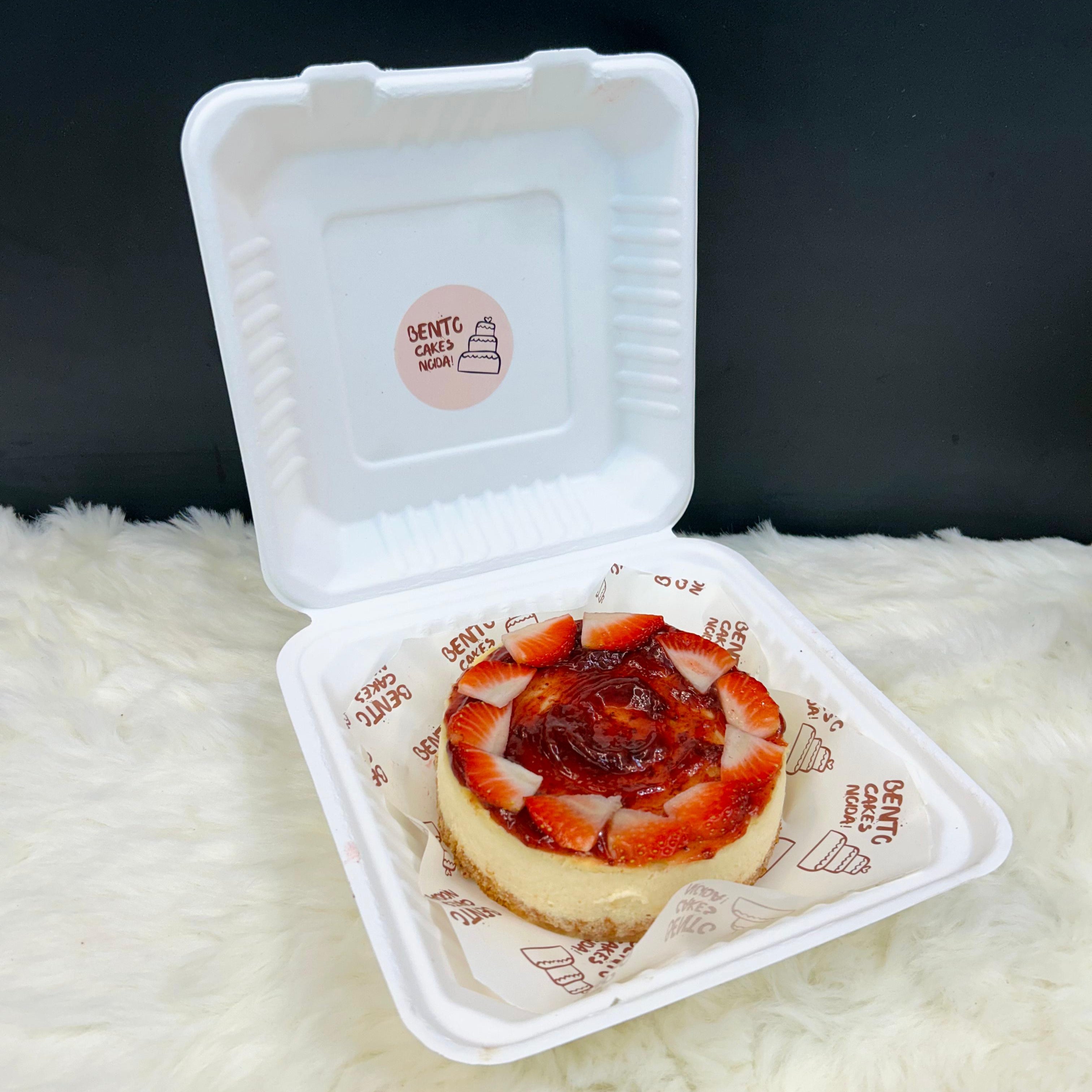 A compact, bento-style strawberry cheesecake topped with fresh strawberry slices and a vibrant glaze, elegantly packaged in a white eco-friendly box on a cozy surface."