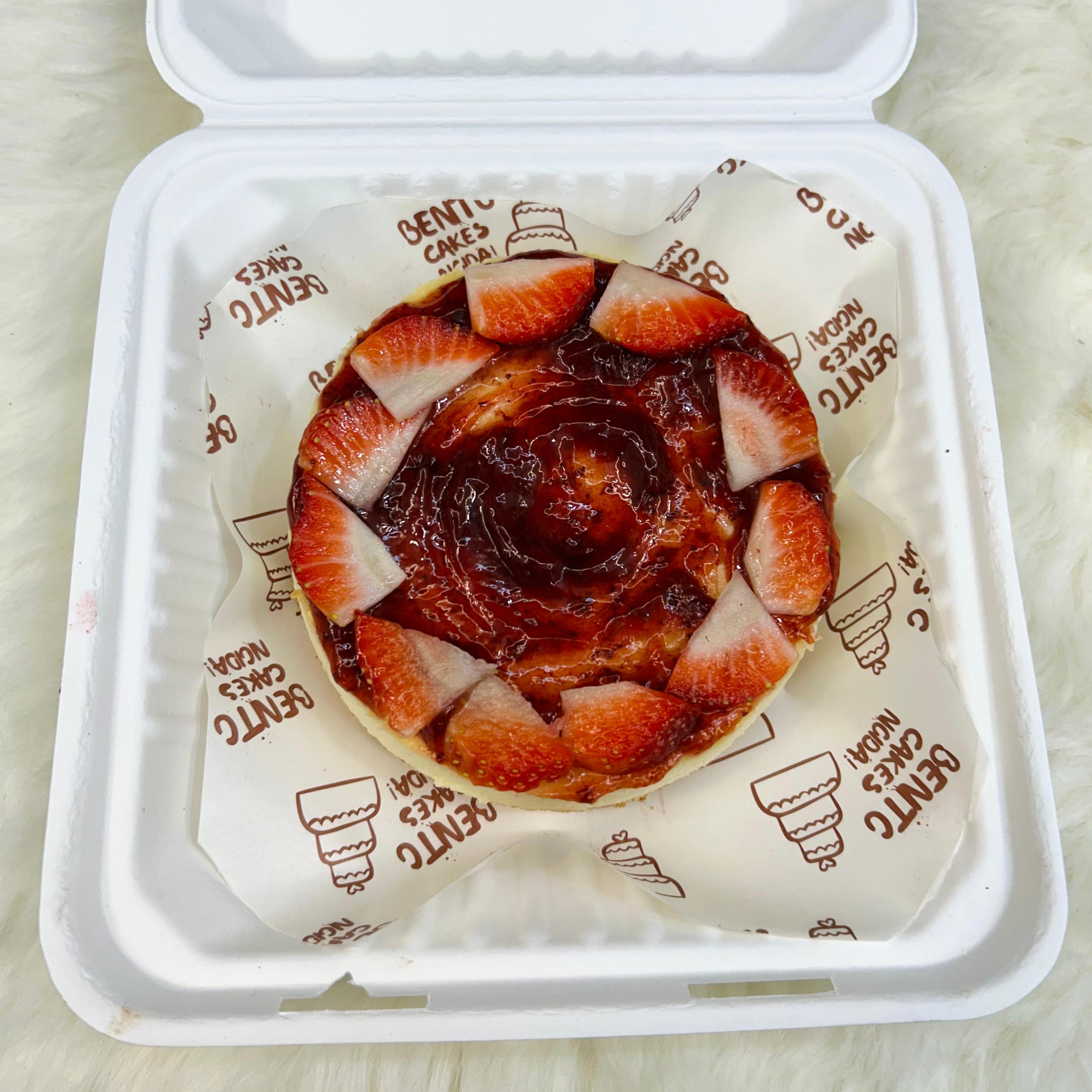 A compact, bento-style strawberry cheesecake topped with fresh strawberry slices and a vibrant glaze, elegantly packaged in a white eco-friendly box on a cozy surface with front view.