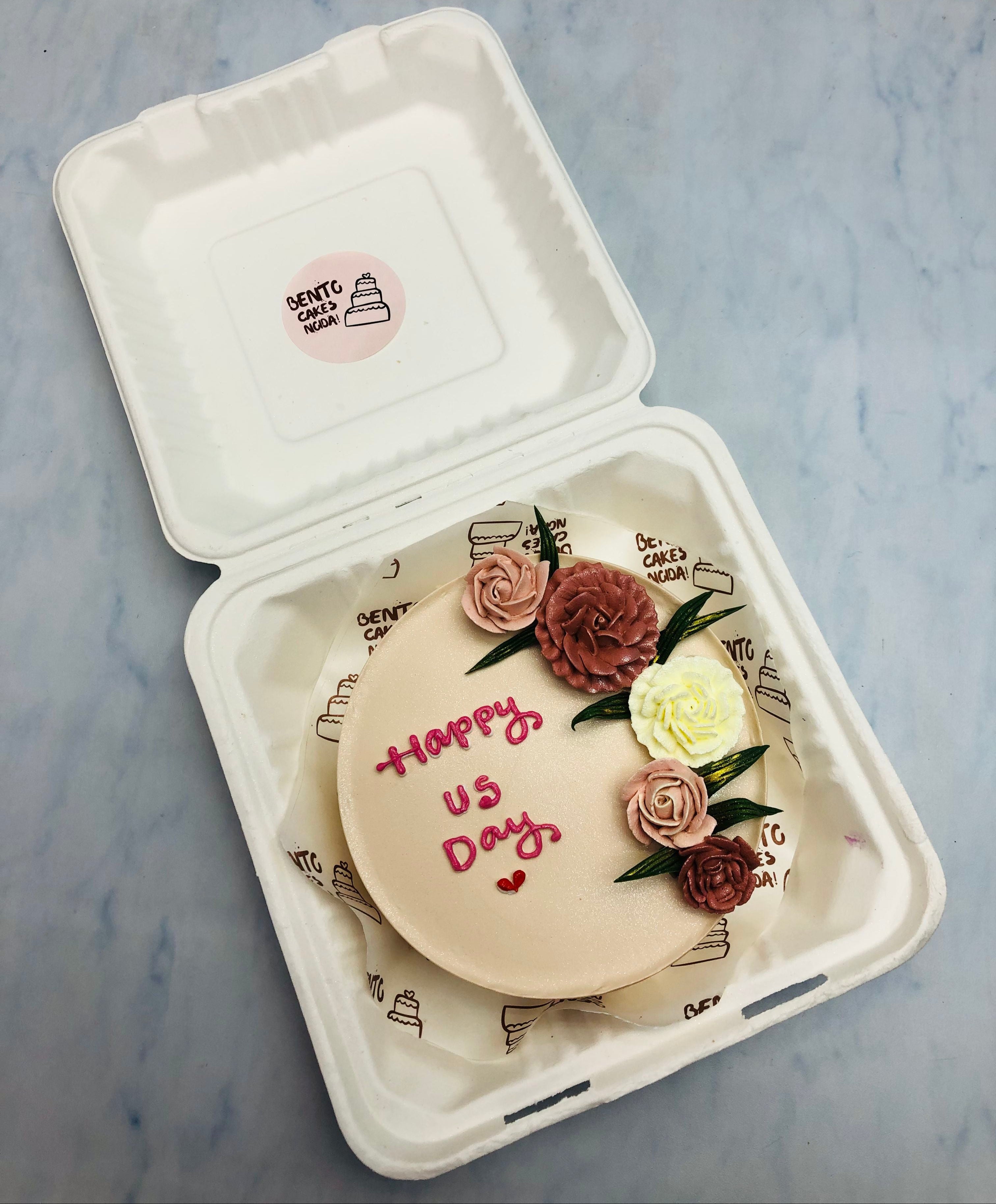 The cake is decorated with five buttercream flowers in shades of pink, brown, and white, along with green leaves. The cake has the message 'Happy Us Day' written in pink icing, accompanied by a small heart.