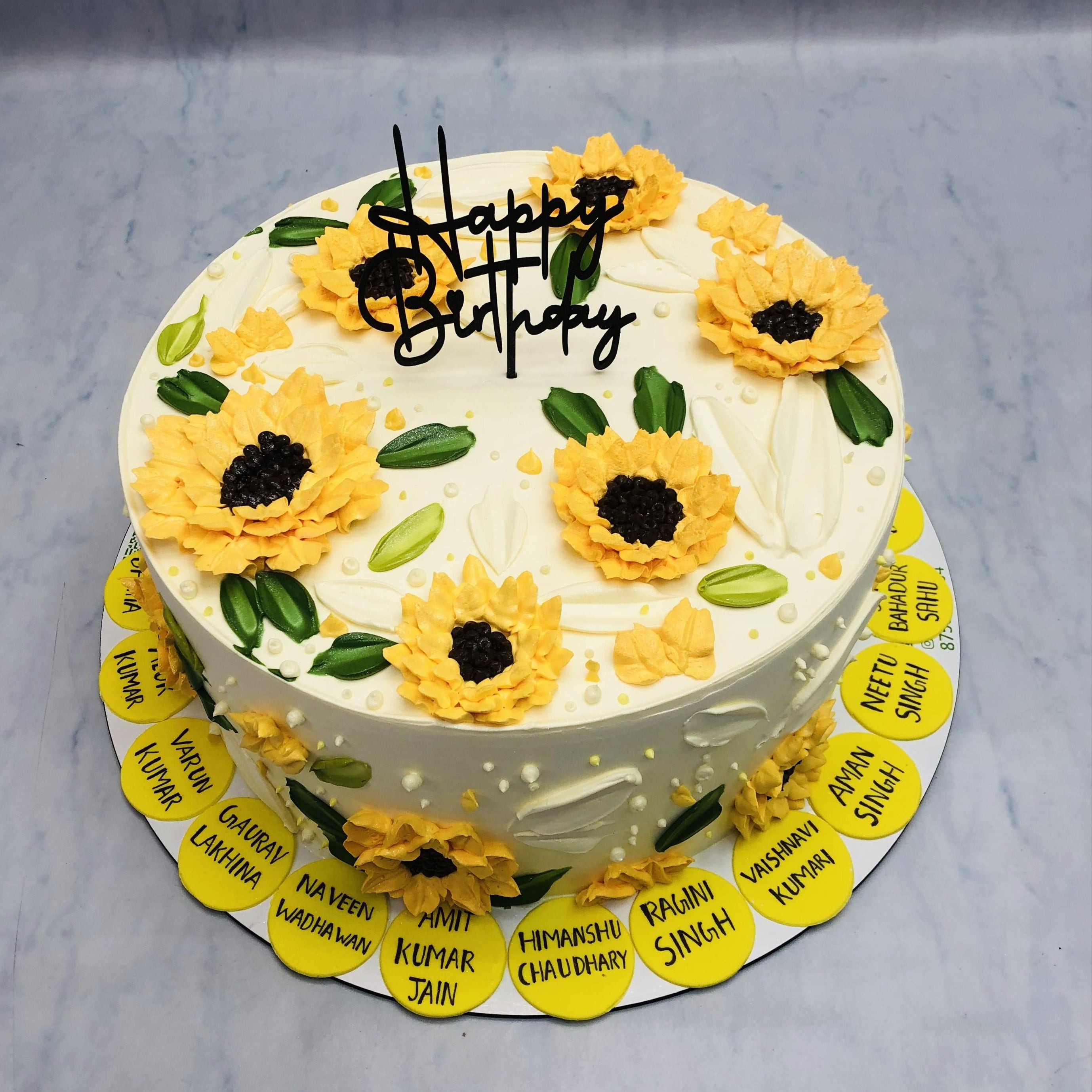 Awhite commercial cake with sunflowers design with name stickers of employes birthday celebration.