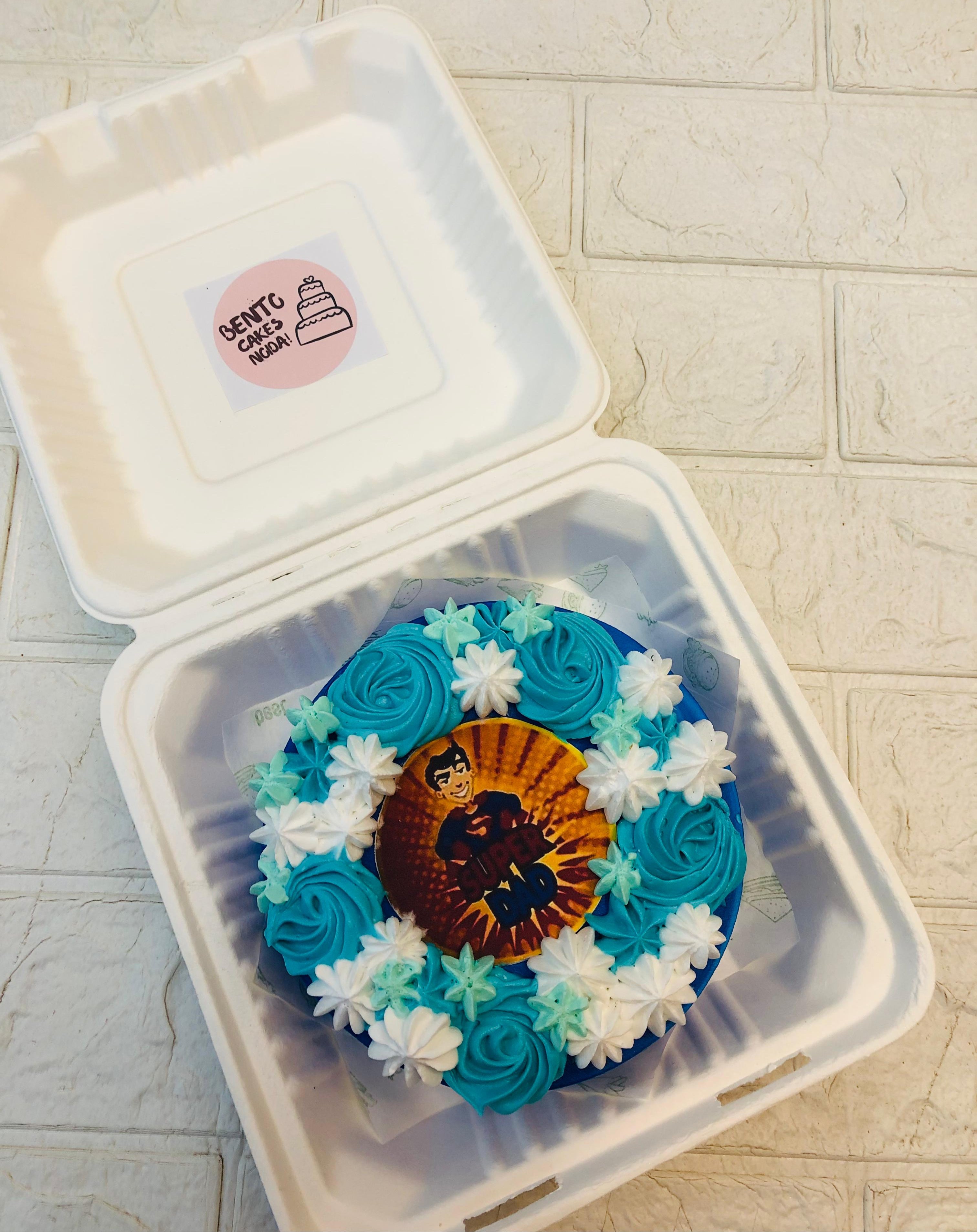 A bento cake decorated with blue and white frosting rosettes and star-shaped decorations, featuring an edible image of a superhero dad with the text "SUPER DAD" in the center, presented in a white bento box.