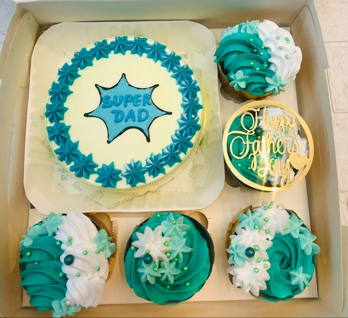 A white bento cake with blue stars design and 5 cubcaks wuth blue and white florals design.