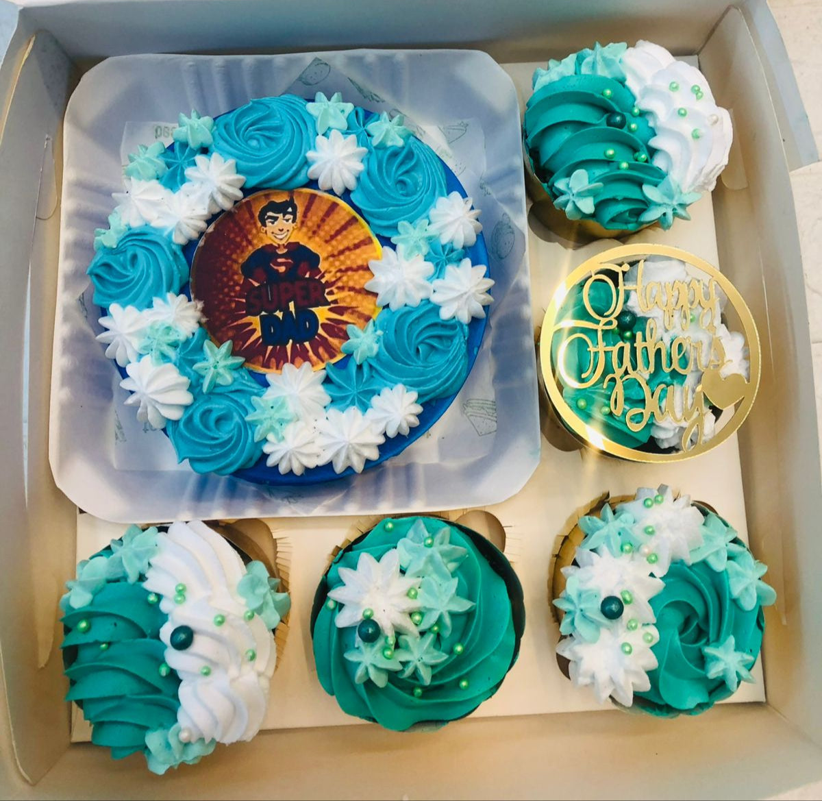 A blue bento cake with blue and white floral design with a super dad cartoon print and 5 cupcakes having blue white floral design.