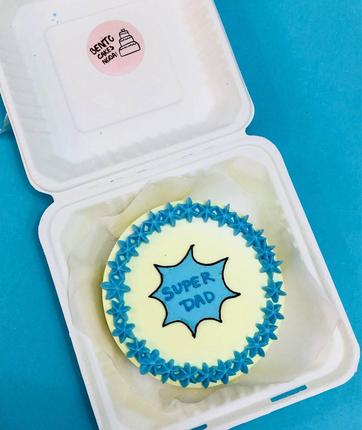 A bento cake in a white box with a blue and white design. The cake has a blue starburst on top with the words "SUPER DAD" written in blue. The cake is bordered with blue star-shaped icing. 