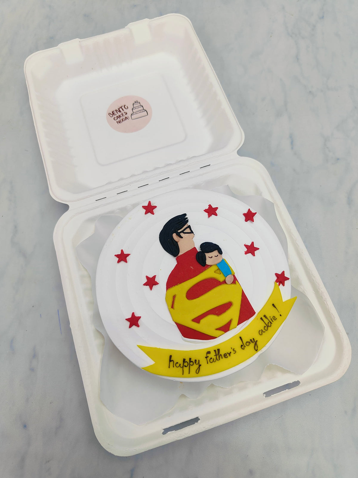 A dad in superman costume with a sleepy baby in hands for fathers day celebration and the cake decorated with little red stars.