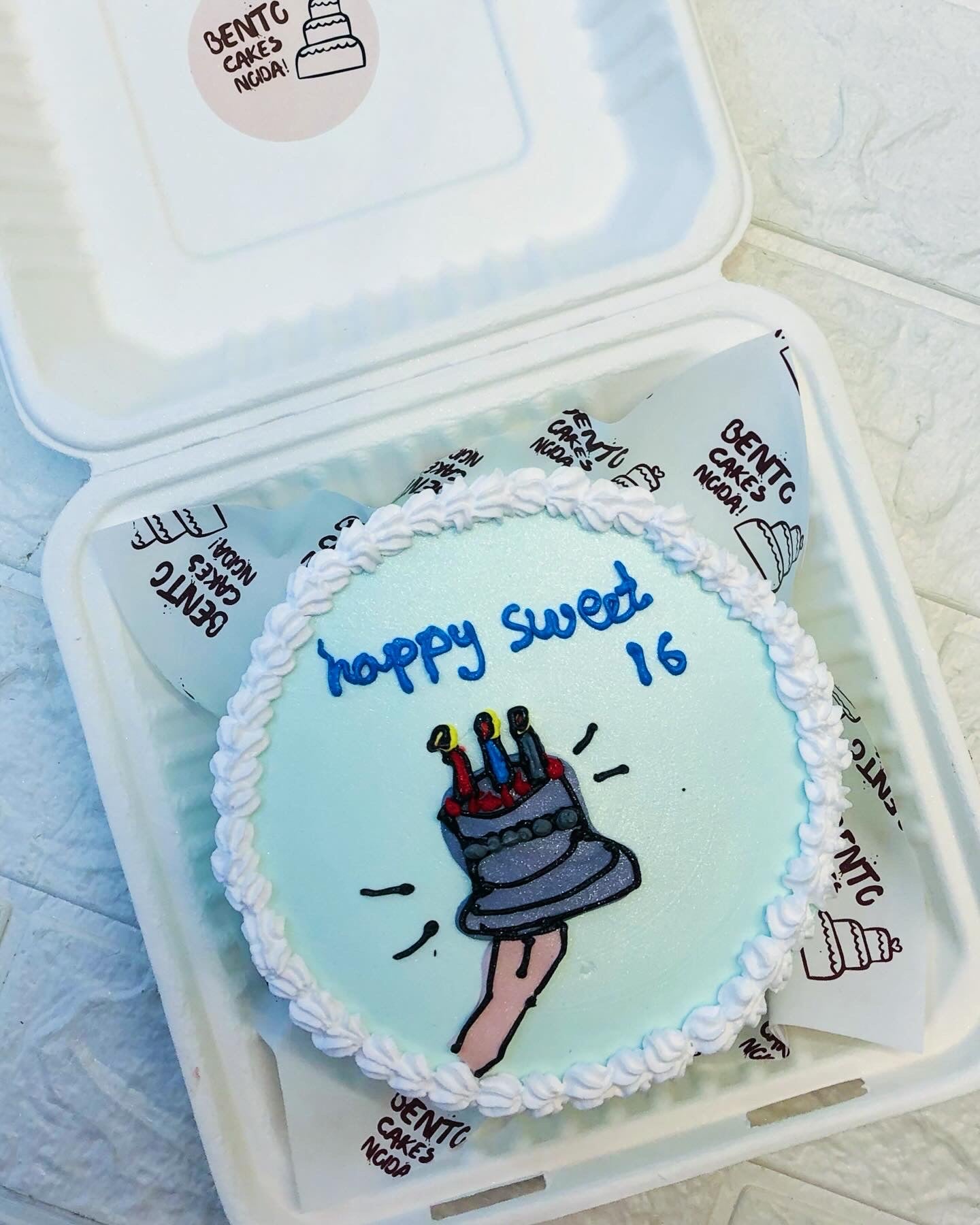 A cool blue bento cake having a design of blue birthday cake in hand and a happy sweet 16 text written.