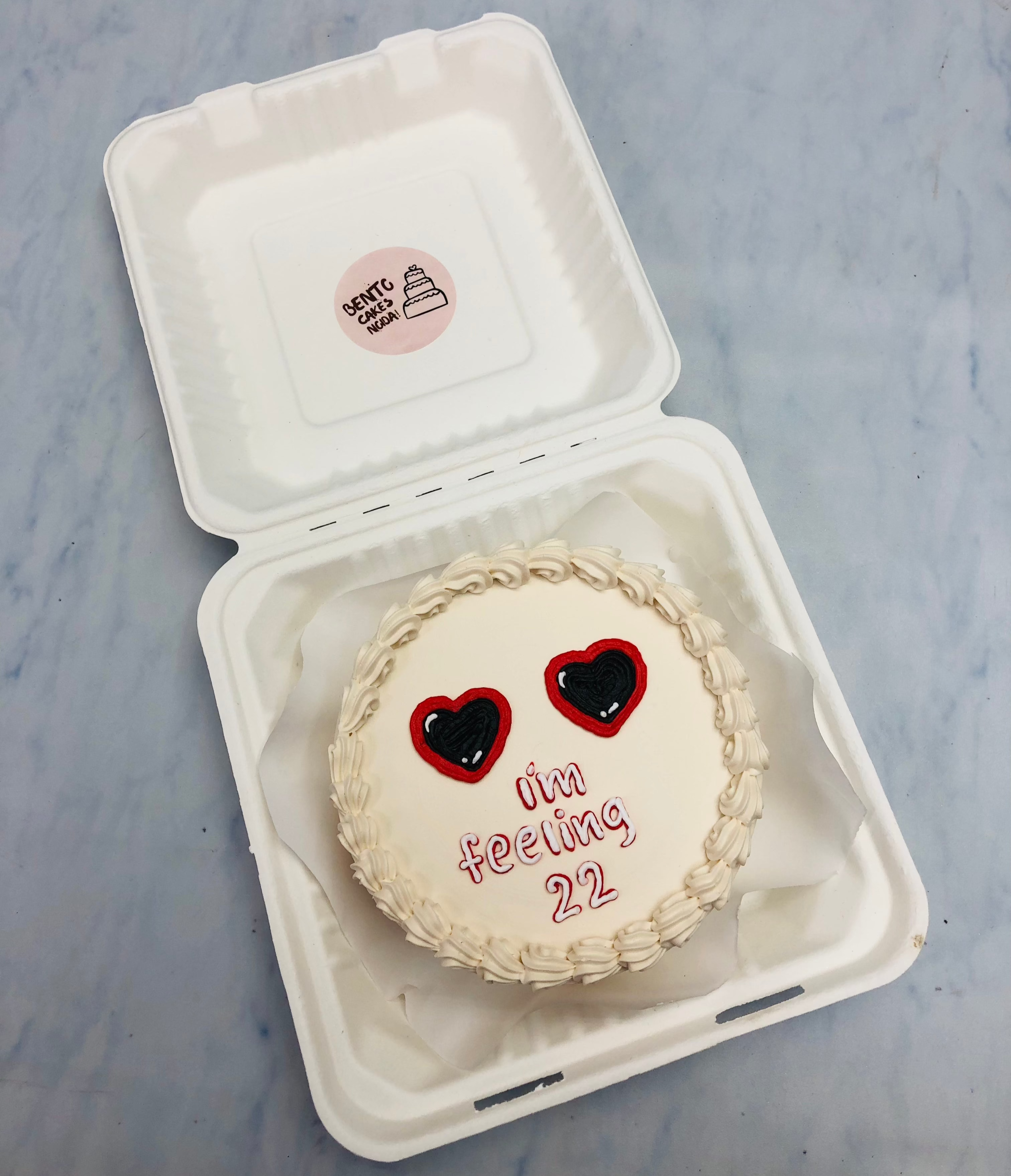 A white bento cake decorated with two heart designs and the text ‘I’m feeling 22’ in red icing.