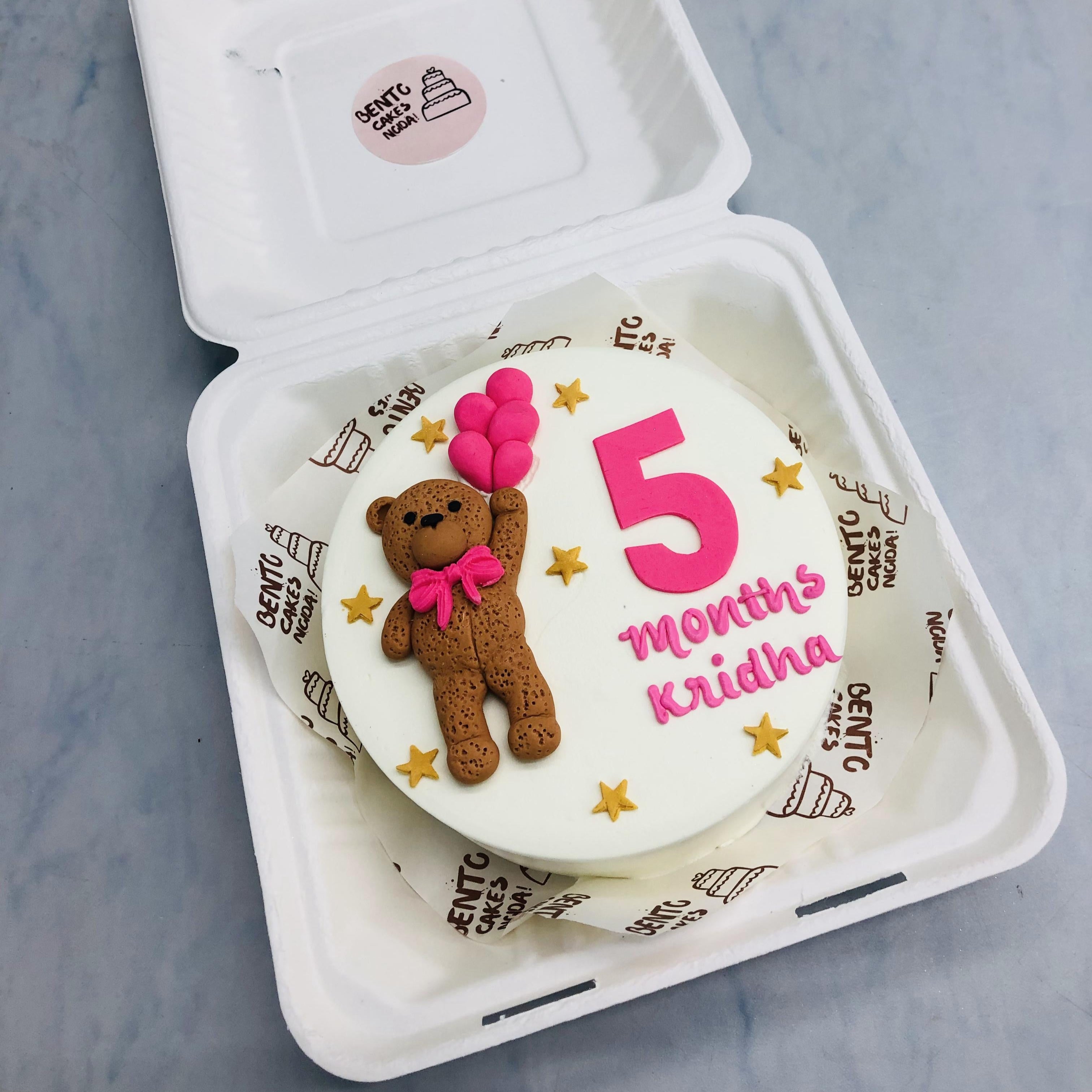 A fondant teddy bear having pink balloon with 5 months and stars on a white bento cake.