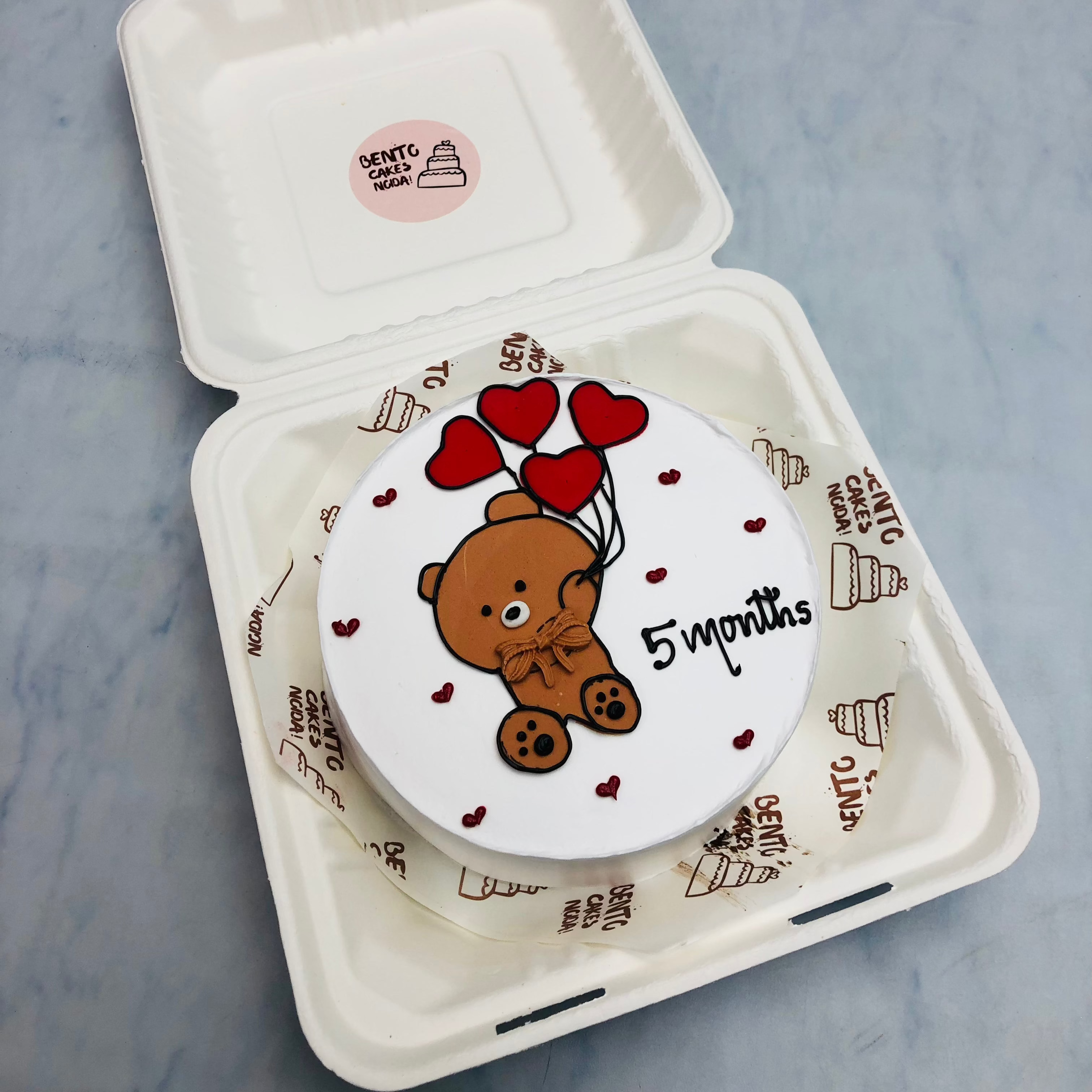A white bento cake with a cute teddy drawing taken red heart shape balloons.
