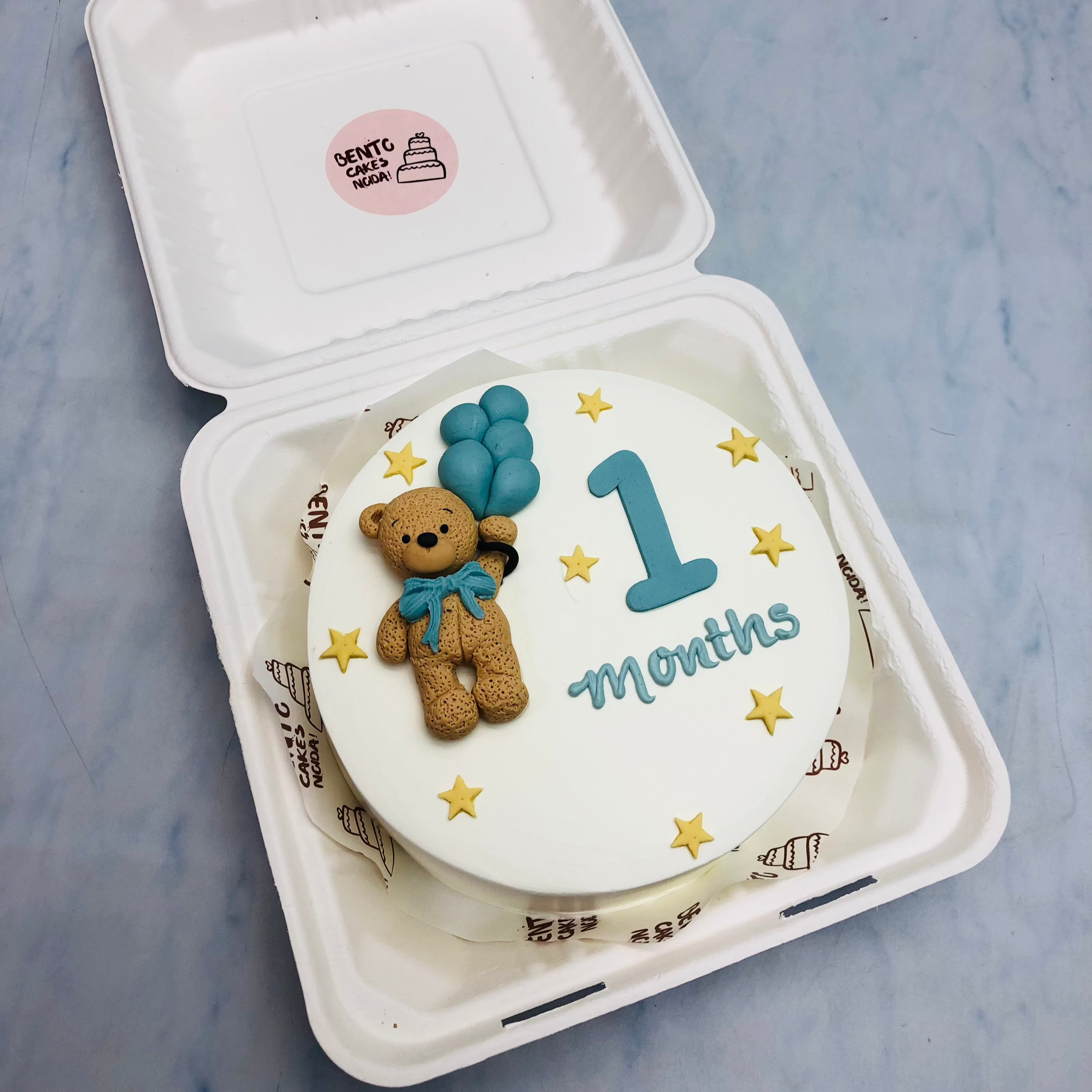 A white cake with a fondant teddy bear design taken blue balloons for 1 month baby.