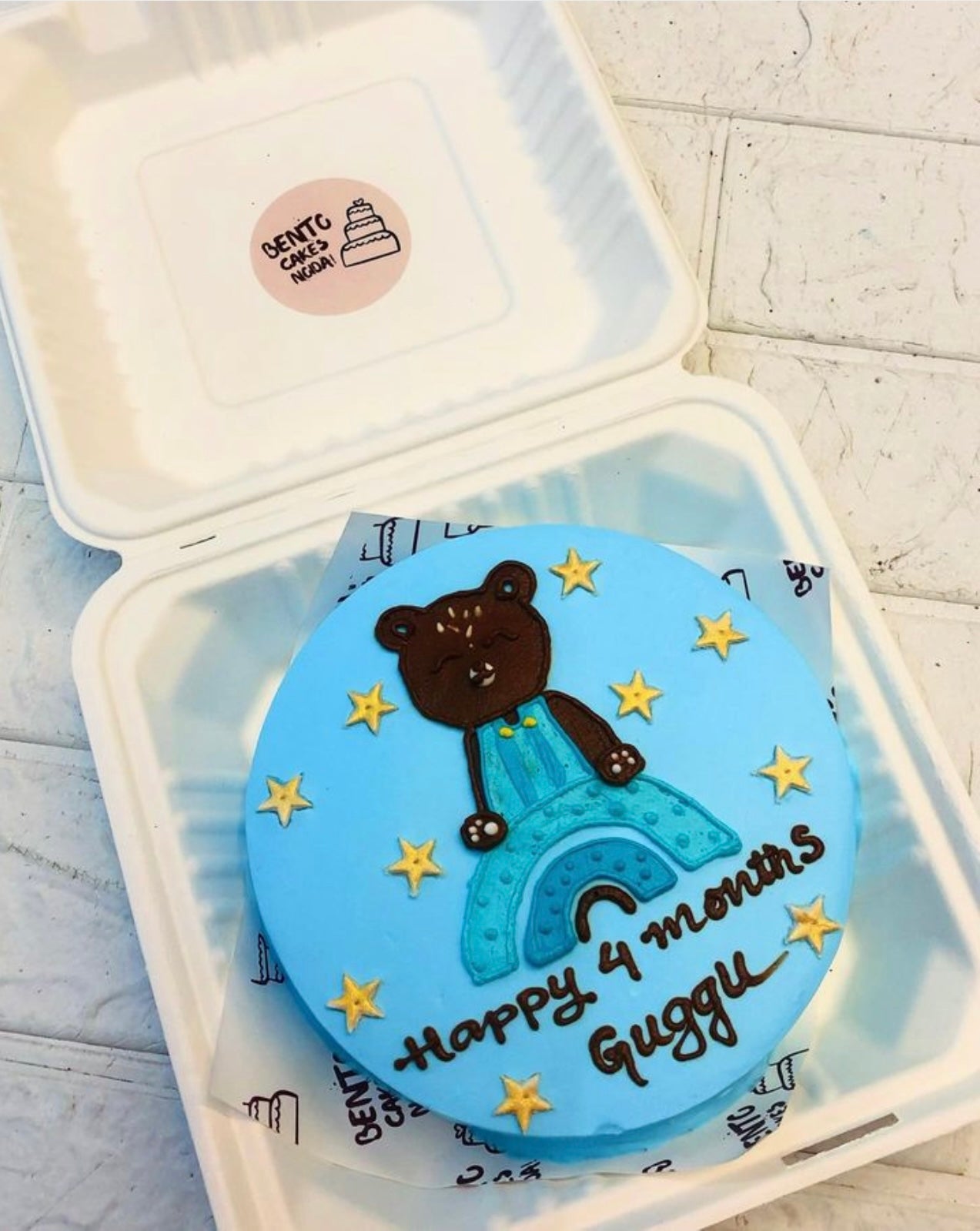 Alight blue cake in a bento box with a blue teddy design on it and the cake filled with golde stars.