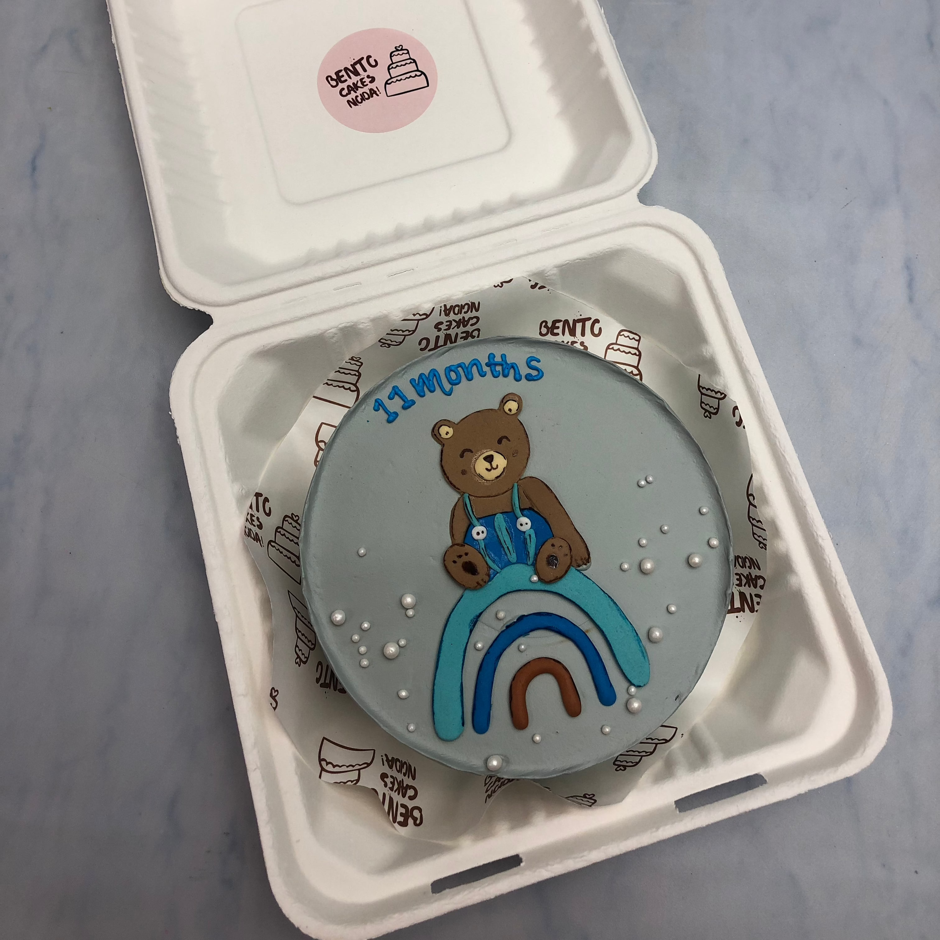A grey color bento cake with a cute teddy sitting on rainbow design.