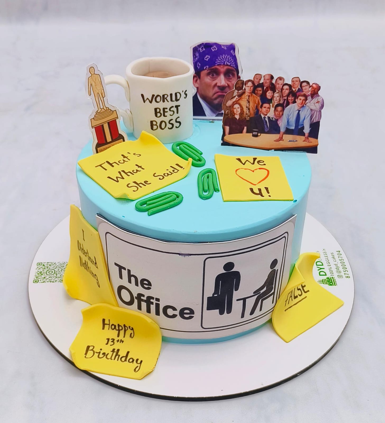 A blue birthday cake decorated with elements inspired by The Office, including a “World’s Best Boss” mug, Dundie Award trophy, sticky notes with quotes from the show, and paper clips.