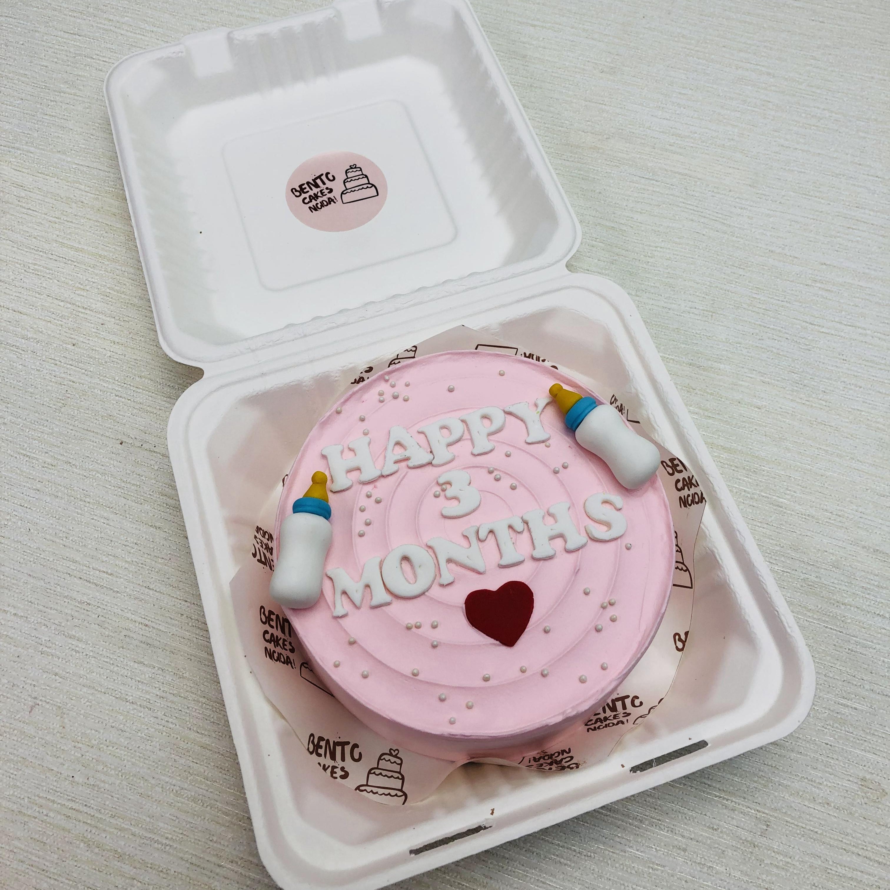 A light pink cake in a bento box having 3 months text and fondant baby feeding bottles.