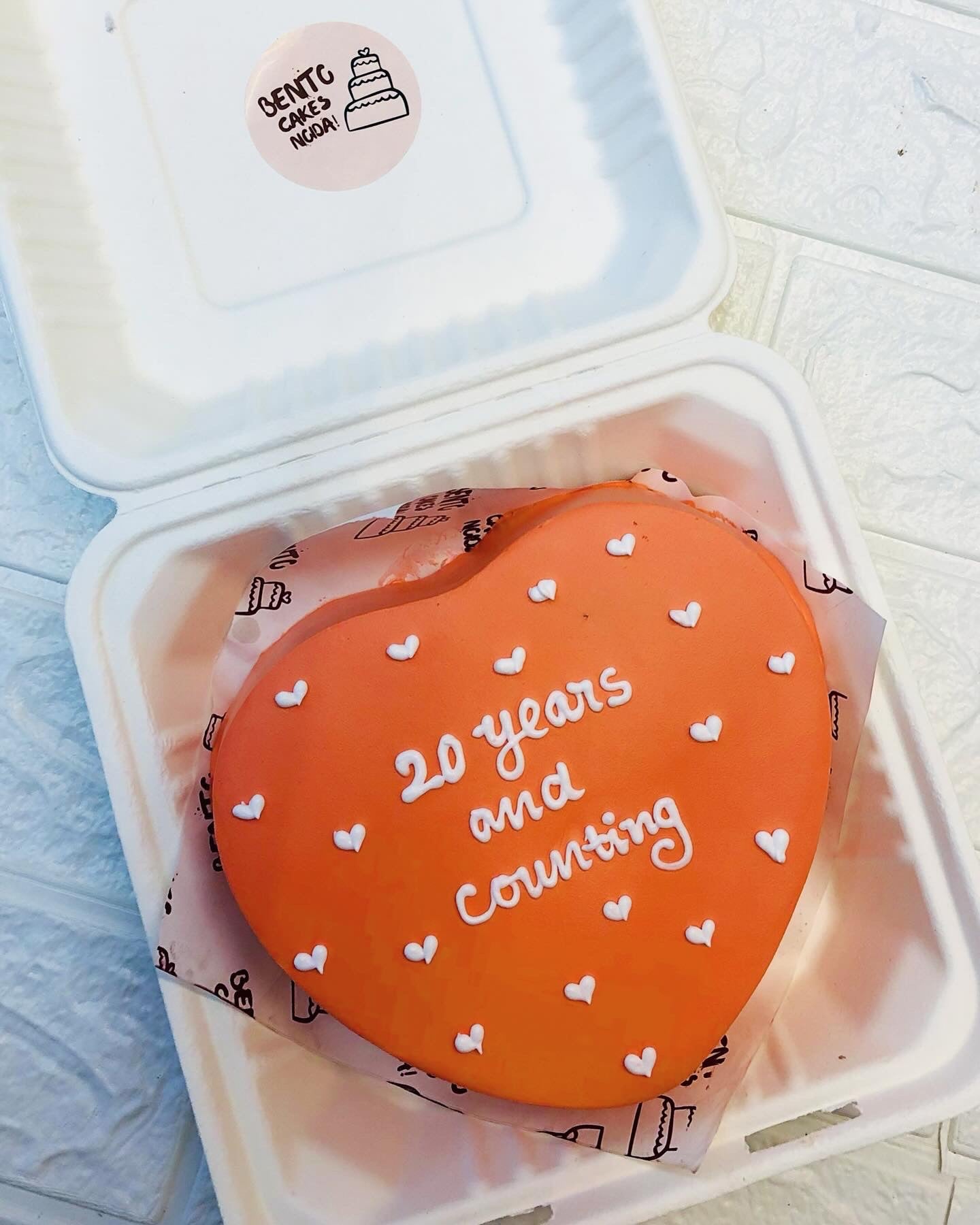 A dark red heart shape cake with white hearts designon it's top and a text "20 years and counting" written with white icing on it.